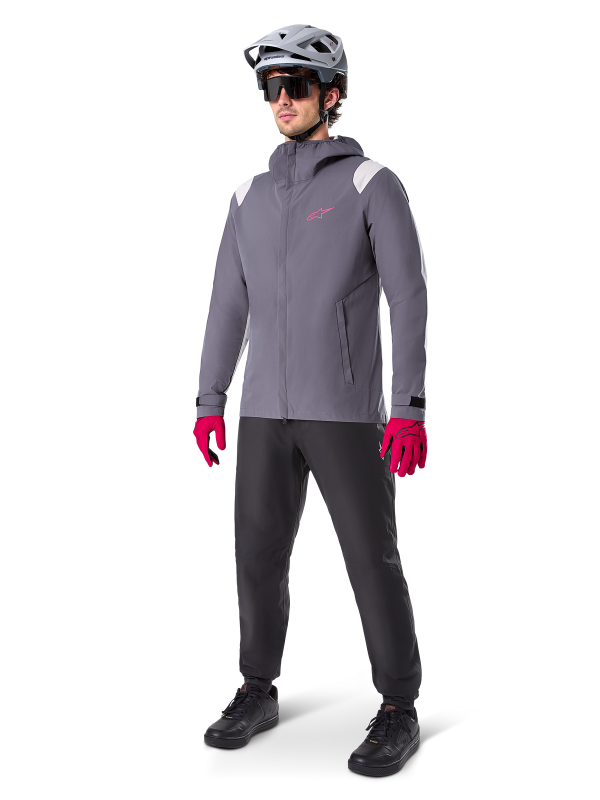 Modelo masculino con la chaqueta impermeable Alpinestars A-Dura en color Dark Titanium, chaqueta gris con capucha, logotipo rosa en el pecho y paneles gris claro en los hombros, combinada con casco de MTB gris, gafas de sol oscuras, guantes rosa vibrante y pantalones negros para ciclismo de montaña.
