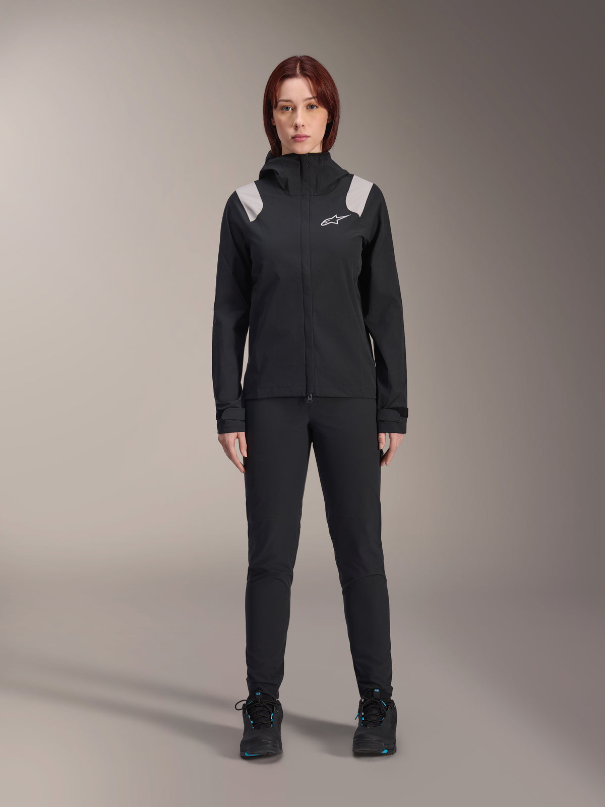 Alpinestars Stella A-Dura Rain Jacket, Women's MTB Rain Jacket, Black with light gray shoulder accents and white logo, worn with matching black pants and shoes, featuring a hood and adjustable cuffs, designed for mountain bike trail riding