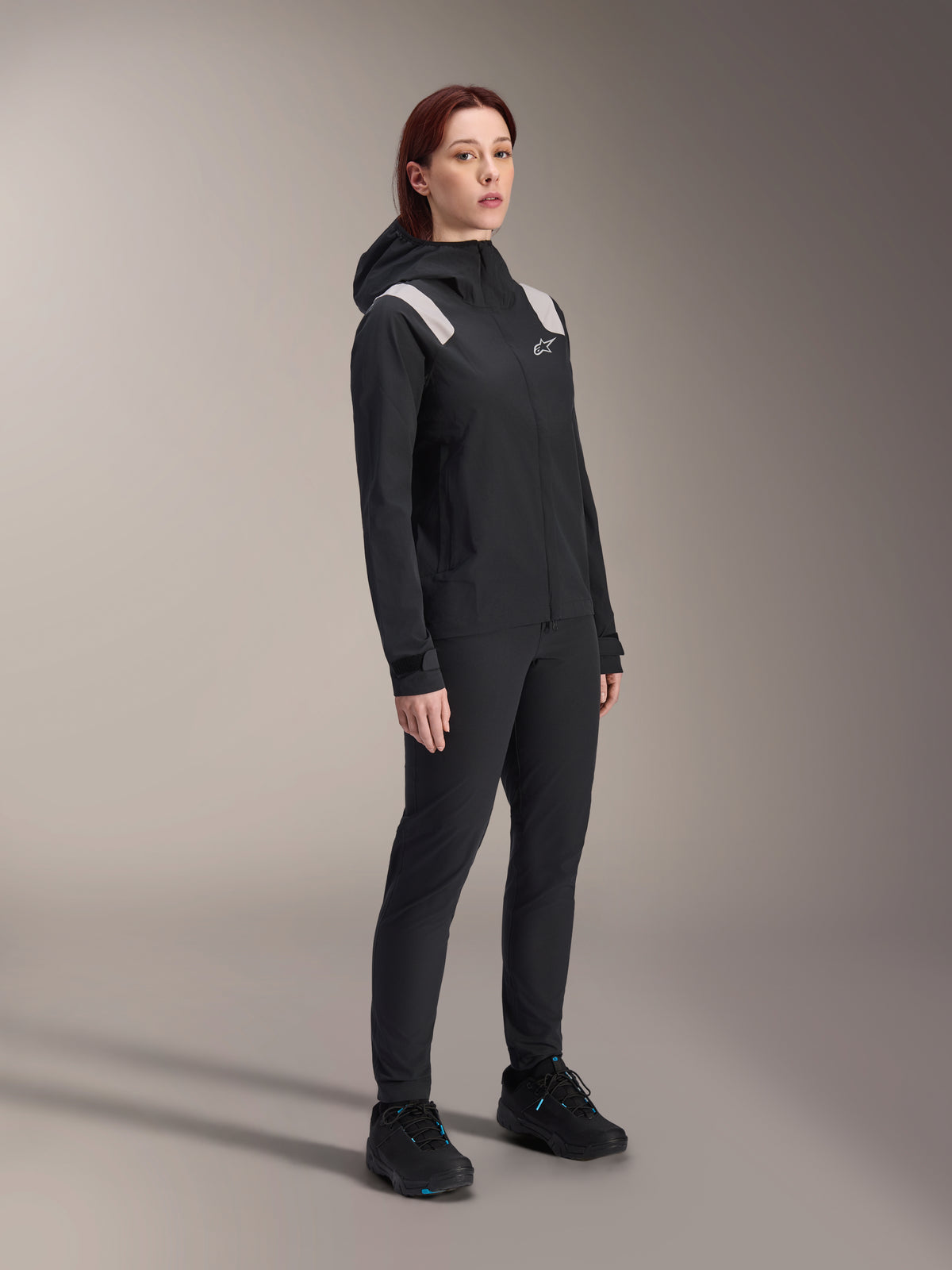 Woman wearing the Alpinestars Stella A-Dura Rain Jacket in Black, 2.5-layer waterproof MTB shell with light grey shoulder accents, matching black pants and cycling shoes, studio portrait for performance cycling gear