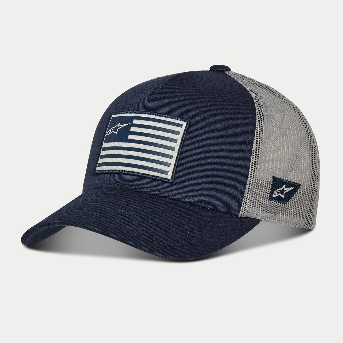 Gorra Alpinestars Flag Snapback, azul marino y gris, visera curva con copa estructurada, parche frontal con bandera estampada y logo lateral bordado, parte trasera de malla para transpirabilidad, accesorio casual inspirado en el automovilismo