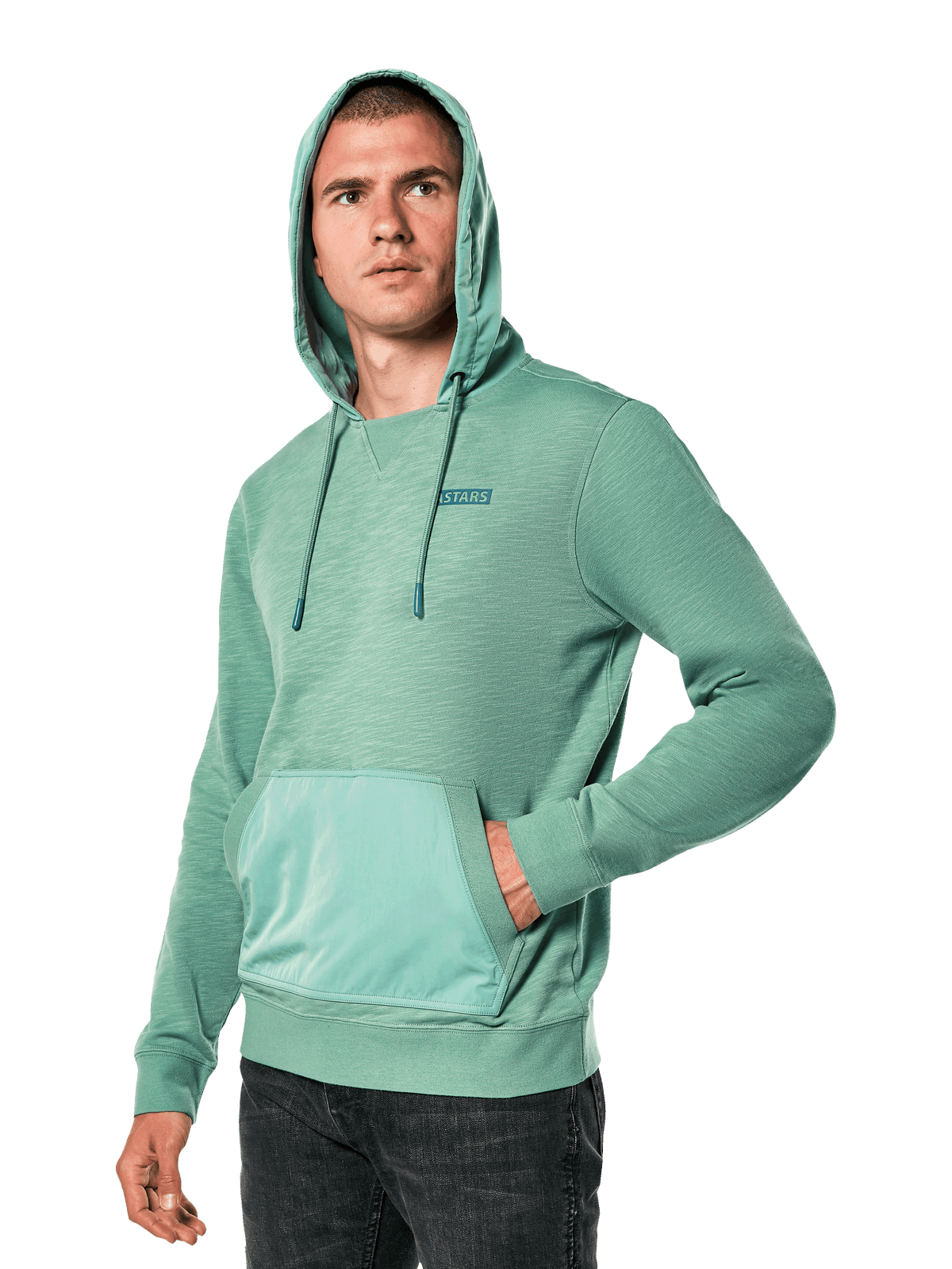 Alpinestars Rooted Hoodie, sudadera de felpa francesa slub verde con capucha superpuesta y bolsillo canguro, pequeño logo STARS en el pecho, lucida por un modelo con la capucha puesta y una mano en el bolsillo, estilo urbano casual inspirado en el motociclismo