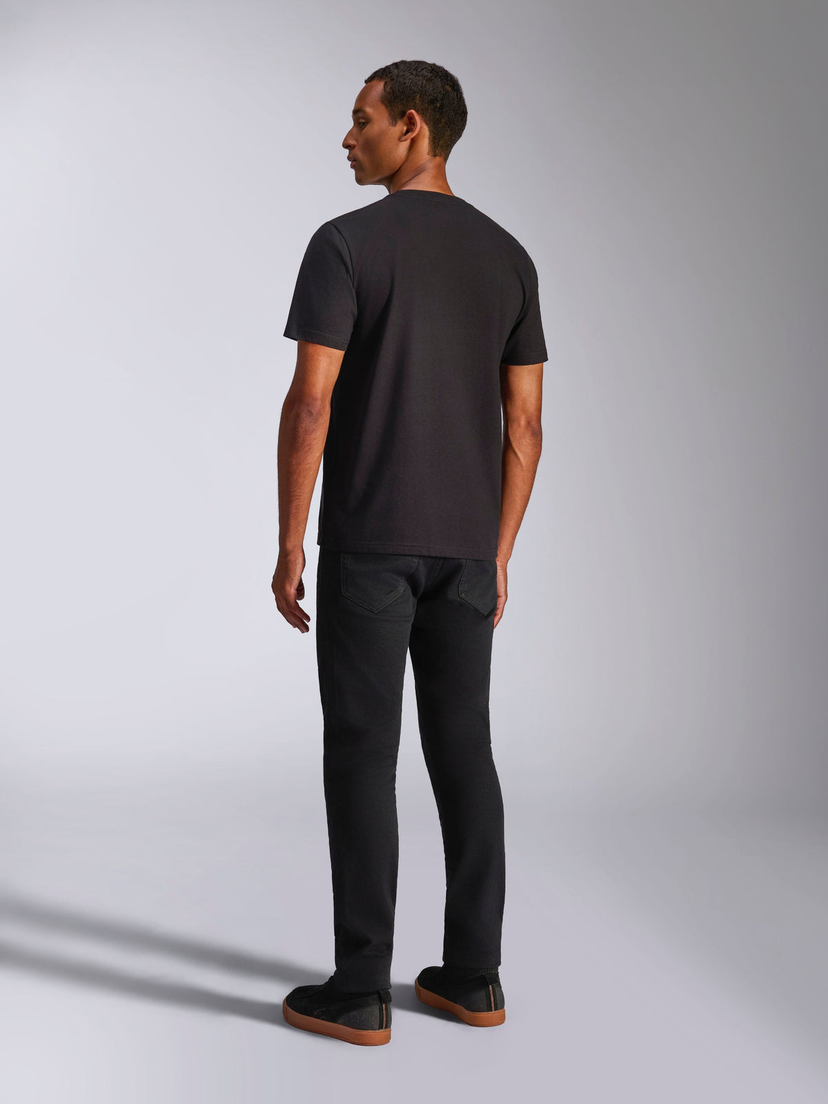 Alpinestars Ageless 2.0 CSF Tee, camiseta de manga corta, negra, vista posterior en un modelo masculino, combinada con pantalones negros y zapatillas con suela de goma, estilo streetwear casual inspirado en el automovilismo