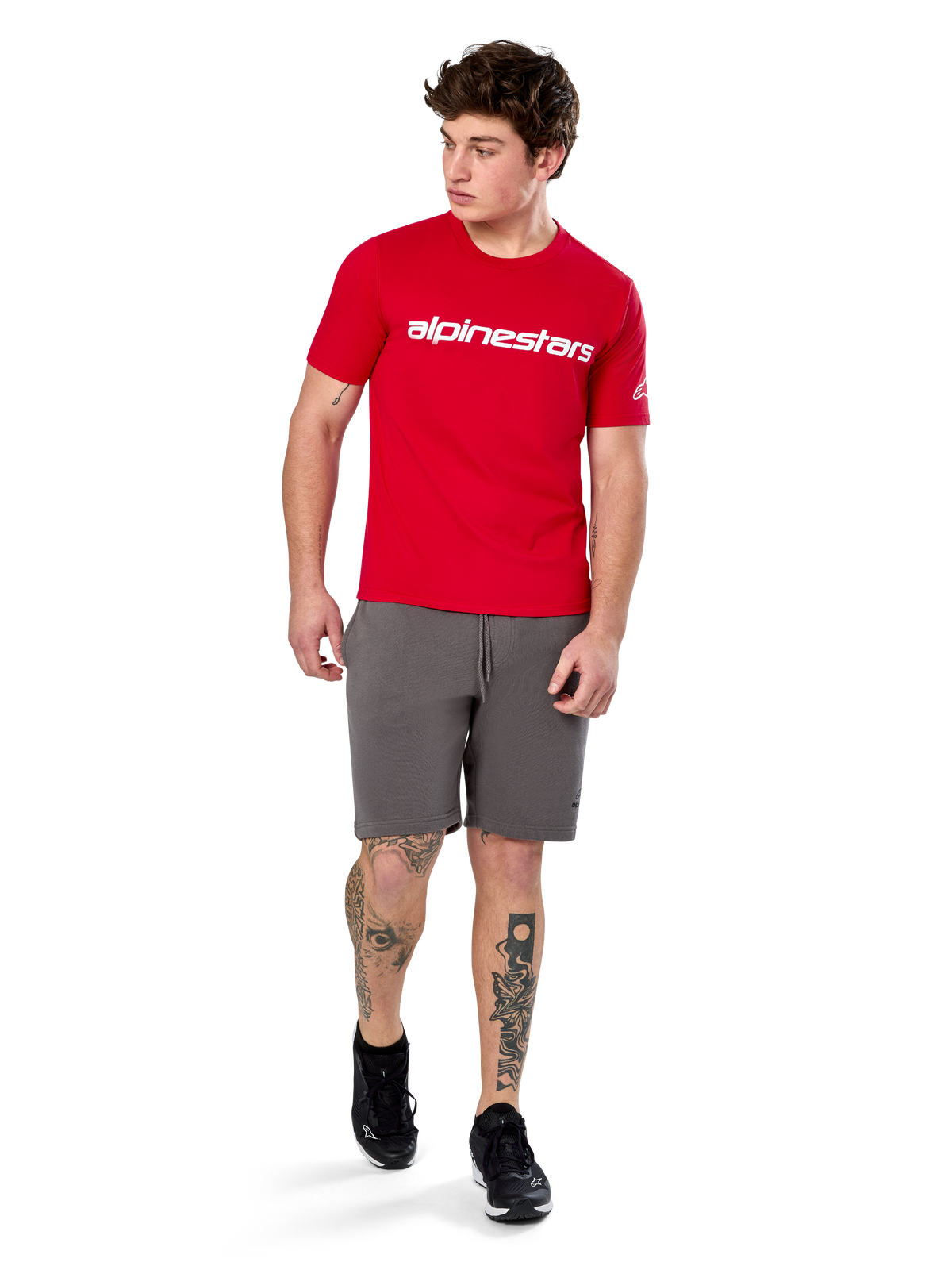 Camiseta Alpinestars Linear Wordmark 2.0 CSF, camiseta de manga corta, roja con logotipo de marca lineal blanco en el pecho, usada por un modelo con pantalones cortos grises y zapatillas negras, ropa urbana casual inspirada en el automovilismo