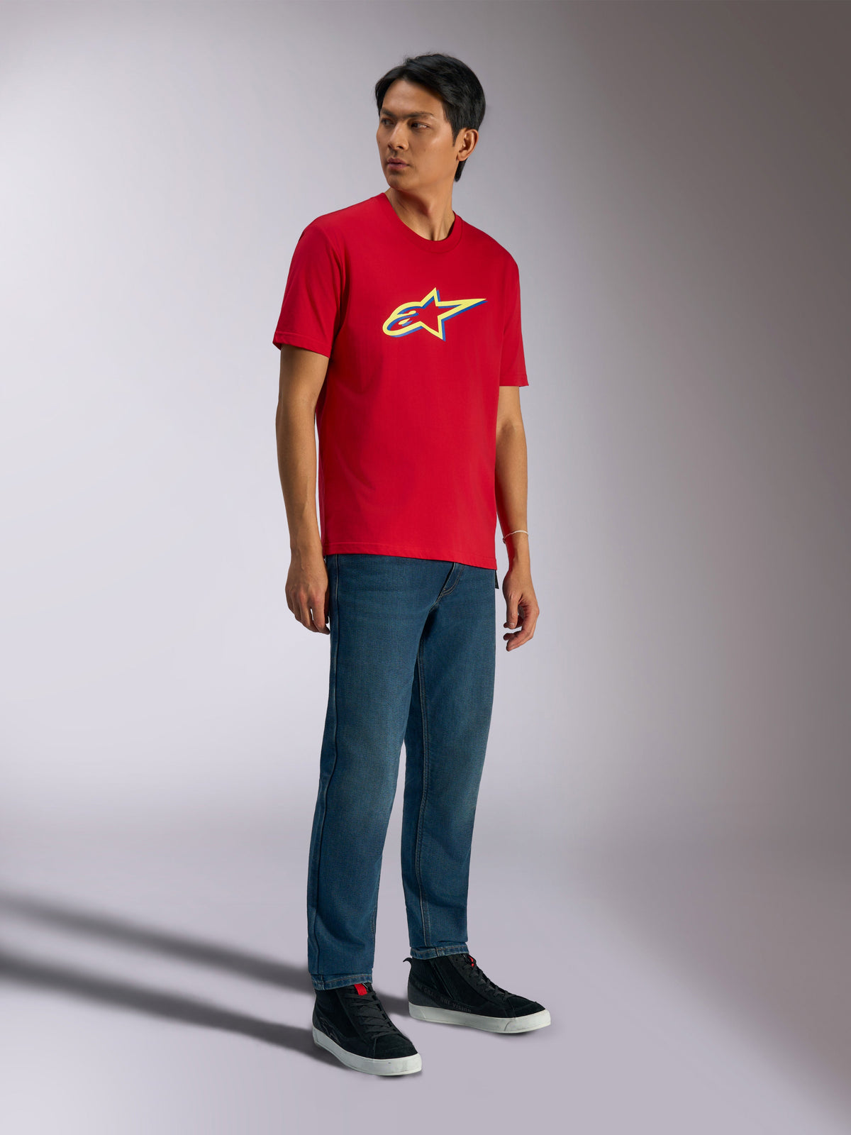 Alpinestars Ageless Shadow CSF Tee, camiseta de manga corta, roja, con el logotipo Ageless Shadow con detalles en amarillo y azul, lucida por un modelo con pantalones vaqueros y zapatillas negras, ropa de calle casual de automovilismo
