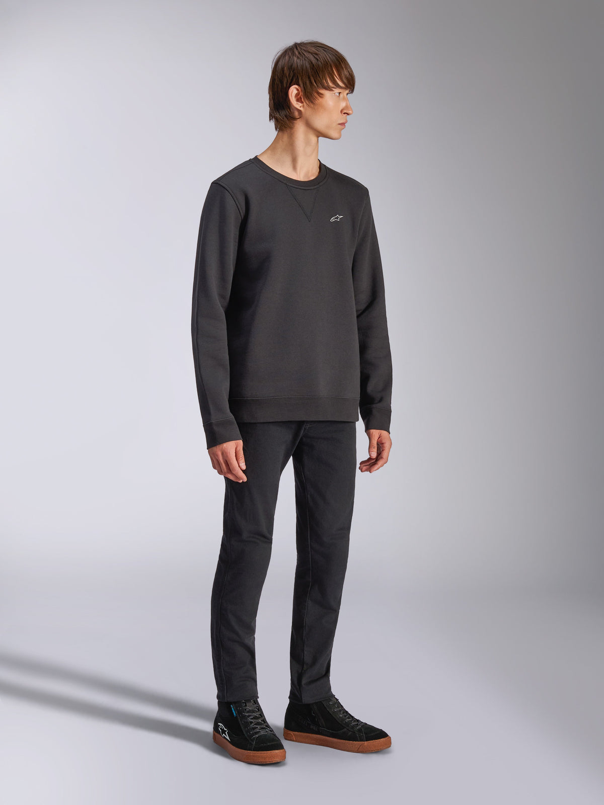 Alpinestars Positive Crew, sudadera de cuello redondo, negra, pequeño logotipo blanco de Alpinestars en el pecho, cuello acanalado con detalle de costura en V, puños y dobladillo acanalados, usada por un modelo con pantalones negros y zapatillas, ropa urbana casual inspirada en el automovilismo