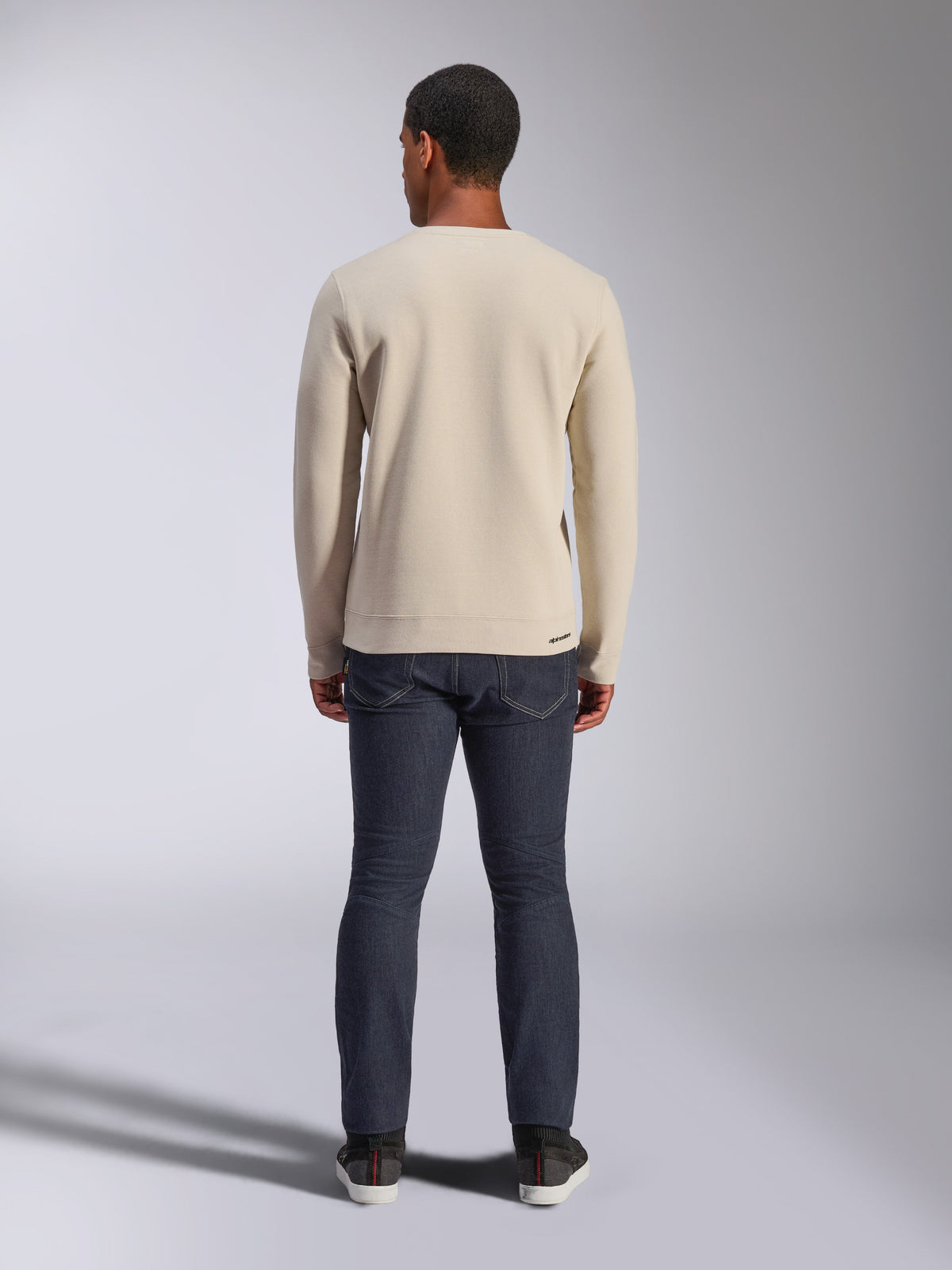 Sudadera Alpinestars Positive Crew, blanco roto, vista trasera de un hombre que viste un jersey beige de cuello redondo con dobladillo y puños de canalé, combinado con vaqueros oscuros, ropa urbana informal inspirada en el automovilismo.