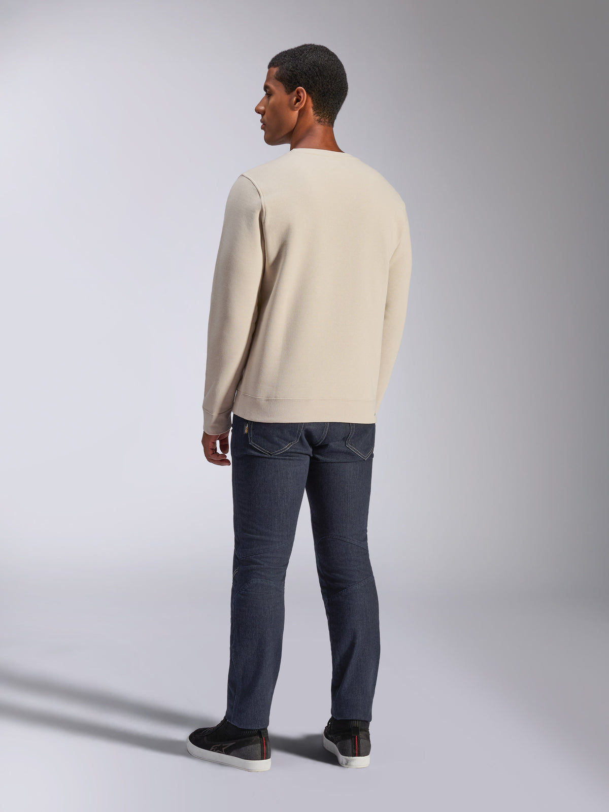 Alpinestars Positive Crew, sudadera de cuello redondo blanco roto, vista trasera en modelo, combinada con vaqueros de denim oscuro y zapatillas negras, estilo urbano casual motorsport, con puños y dobladillo acanalados
