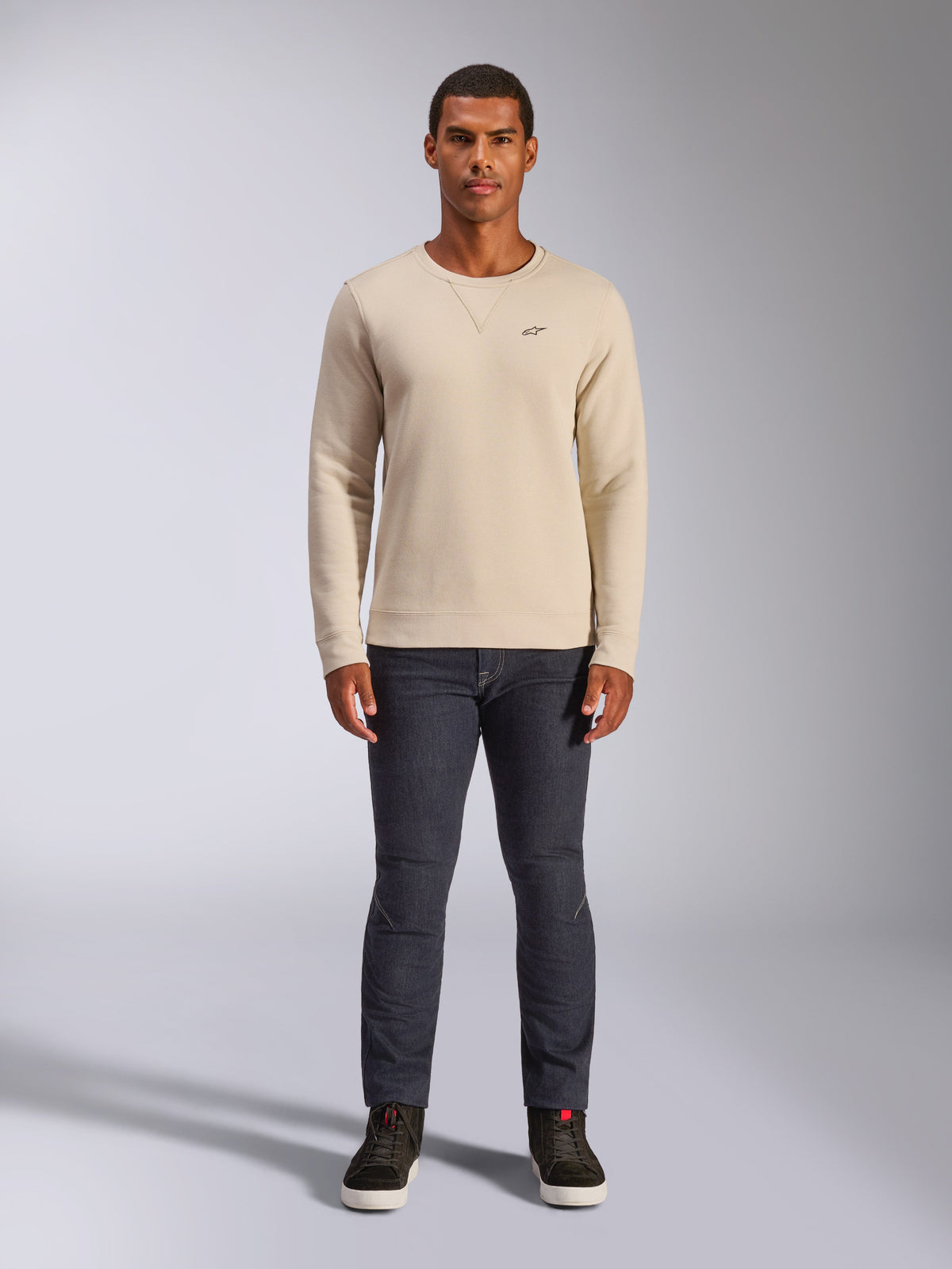 Alpinestars Positive Crew, sudadera de cuello redondo en color blanco roto, pequeño logotipo negro en el pecho, puños y dobladillo acanalados, costura triangular en el cuello, estilo casual motorsport streetwear combinada con vaqueros oscuros