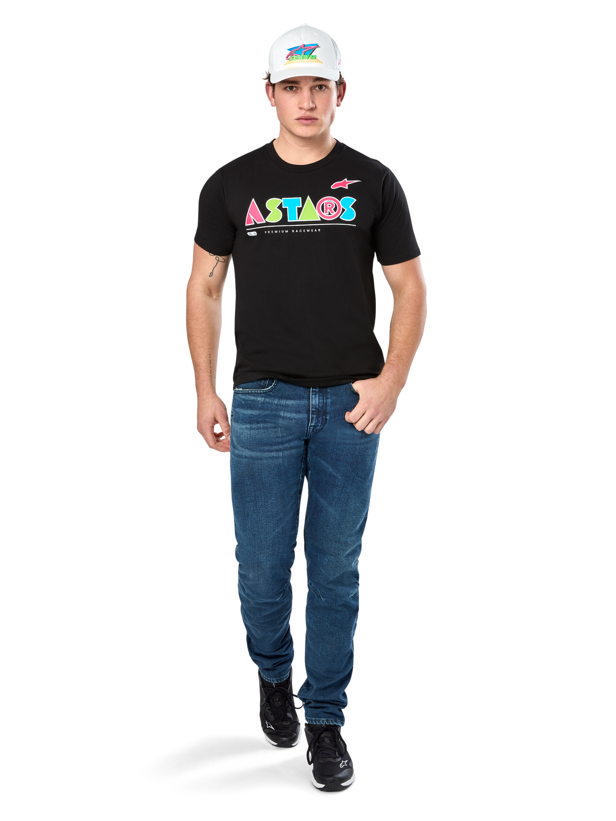 Modelo masculino vistiendo la camiseta Alpinestars Cereal CSF, camiseta negra de manga corta con un colorido logo geométrico de ASTARS, combinada con pantalones vaqueros azules y gorra blanca, estilo streetwear casual inspirado en el automovilismo.