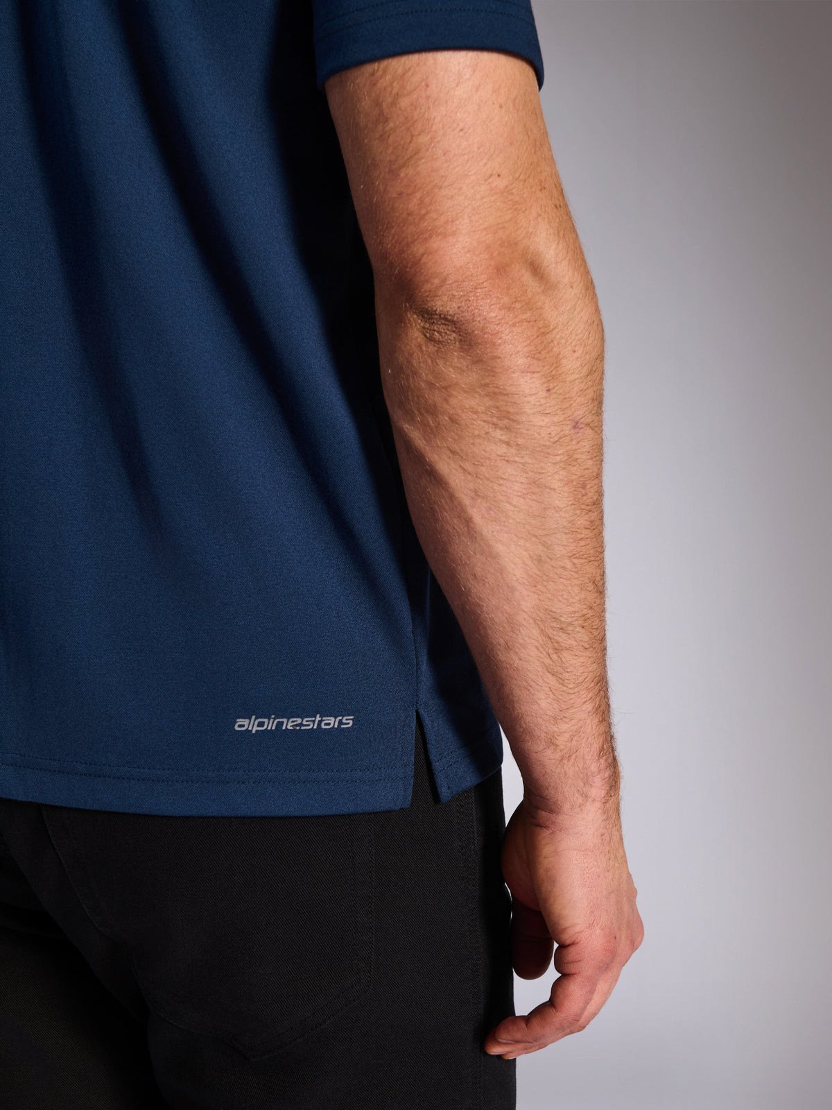 Alpinestars Impetus Sport Polo, azul marino, primer plano del dobladillo inferior que muestra la marca denominativa Alpinestars y la abertura lateral, textura de punto de malla simulada, ropa deportiva casual de automovilismo