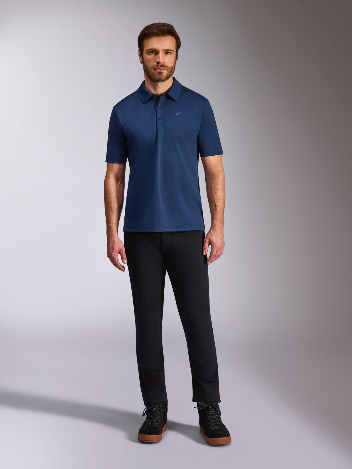 Alpinestars Impetus Sport Polo, polo de manga corta, azul marino, pequeño logo Astars plateado en el pecho, tapeta de tres botones, usado por un modelo con pantalones negros y botas, ropa deportiva casual inspirada en el motorsport