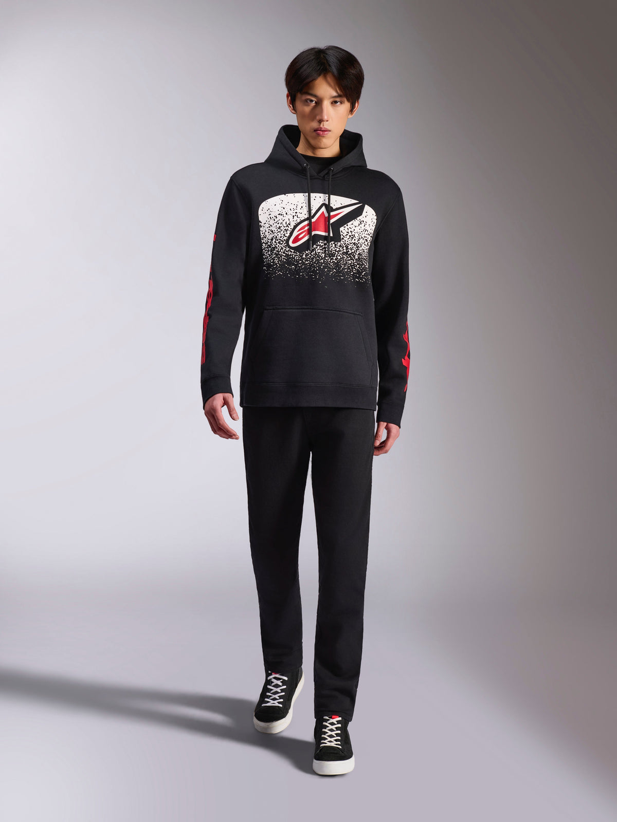 Sudadera con capucha Alpinestars Obsessed, sudadera negra tipo pullover, logotipo grande de Astars en blanco y rojo con degradado MX, logotipos rojos en las mangas, modelo con conjunto casual totalmente negro, ropa lifestyle inspirada en el motociclismo