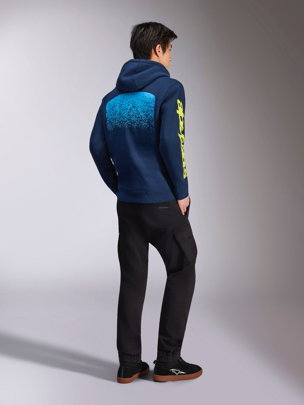 Sudadera con capucha Alpinestars Obsessed, sudadera casual azul marino, vista trasera que muestra el gráfico de degradado MX en azul claro y el logotipo amarillo de Alpinestars en la manga, combinada con pantalones negros y zapatillas