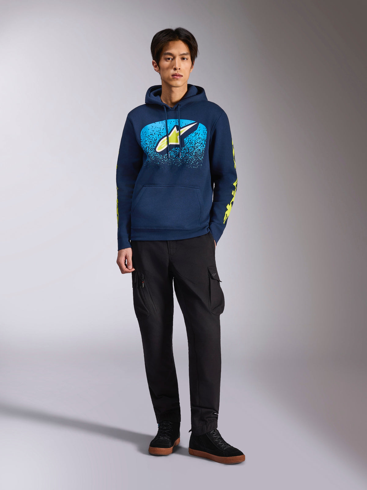Sudadera con capucha Alpinestars Obsessed, sudadera azul marino tipo pullover, logo frontal de Astars con degradado MX azul y estrella amarillo lima, logotipos de Alpinestars en las mangas en amarillo lima, usada por un modelo con pantalones cargo negros y zapatillas negras, ropa urbana informal inspirada en el automovilismo