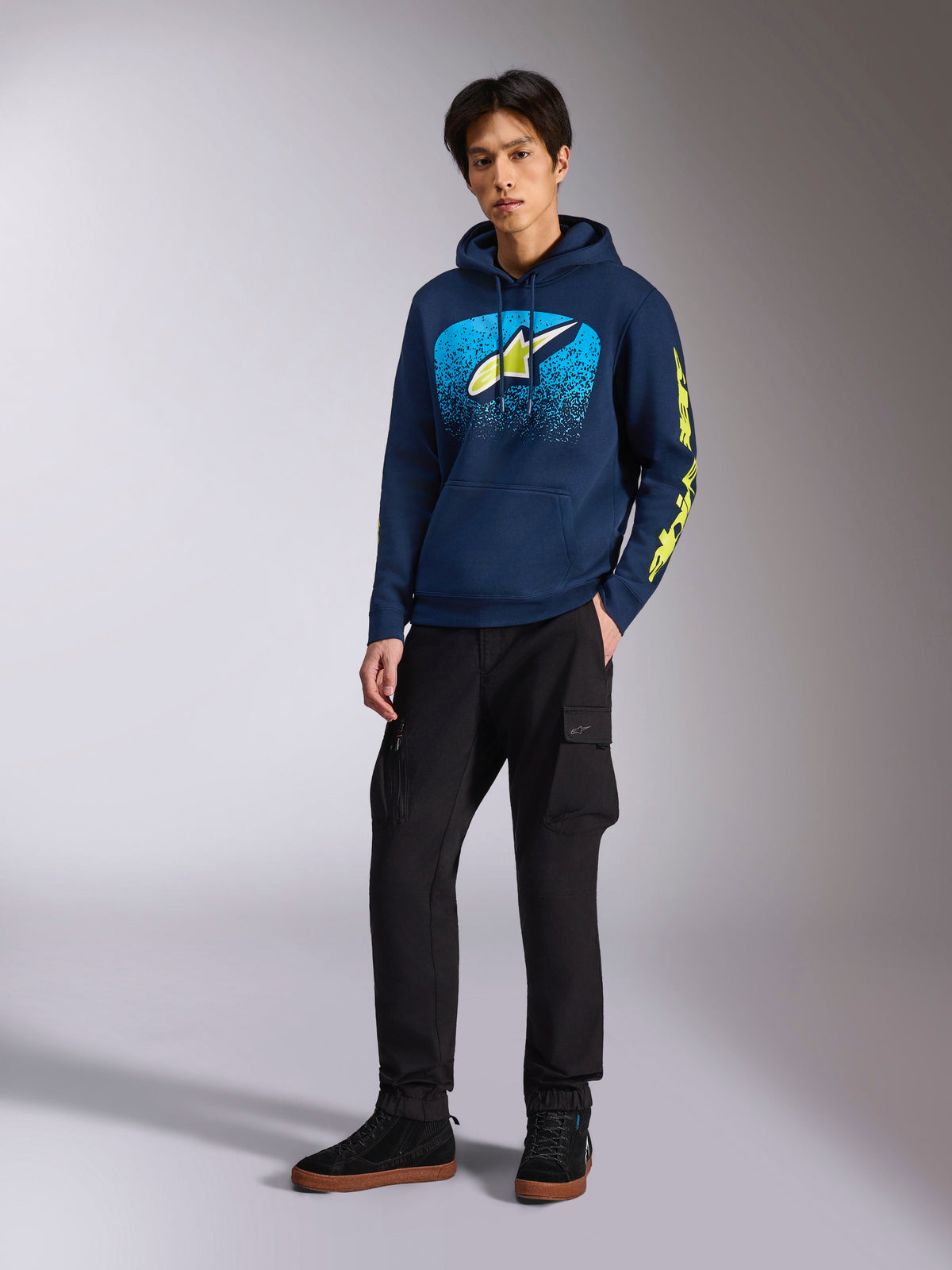 Sudadera con capucha Alpinestars Obsessed en azul marino, sudadera casual de forro polar con gráfico frontal en azul claro y amarillo lima, logotipos de Alpinestars en amarillo lima en la manga, lucida por un modelo con pantalones cargo negros y zapatillas negras, estilo streetwear inspirado en el motor