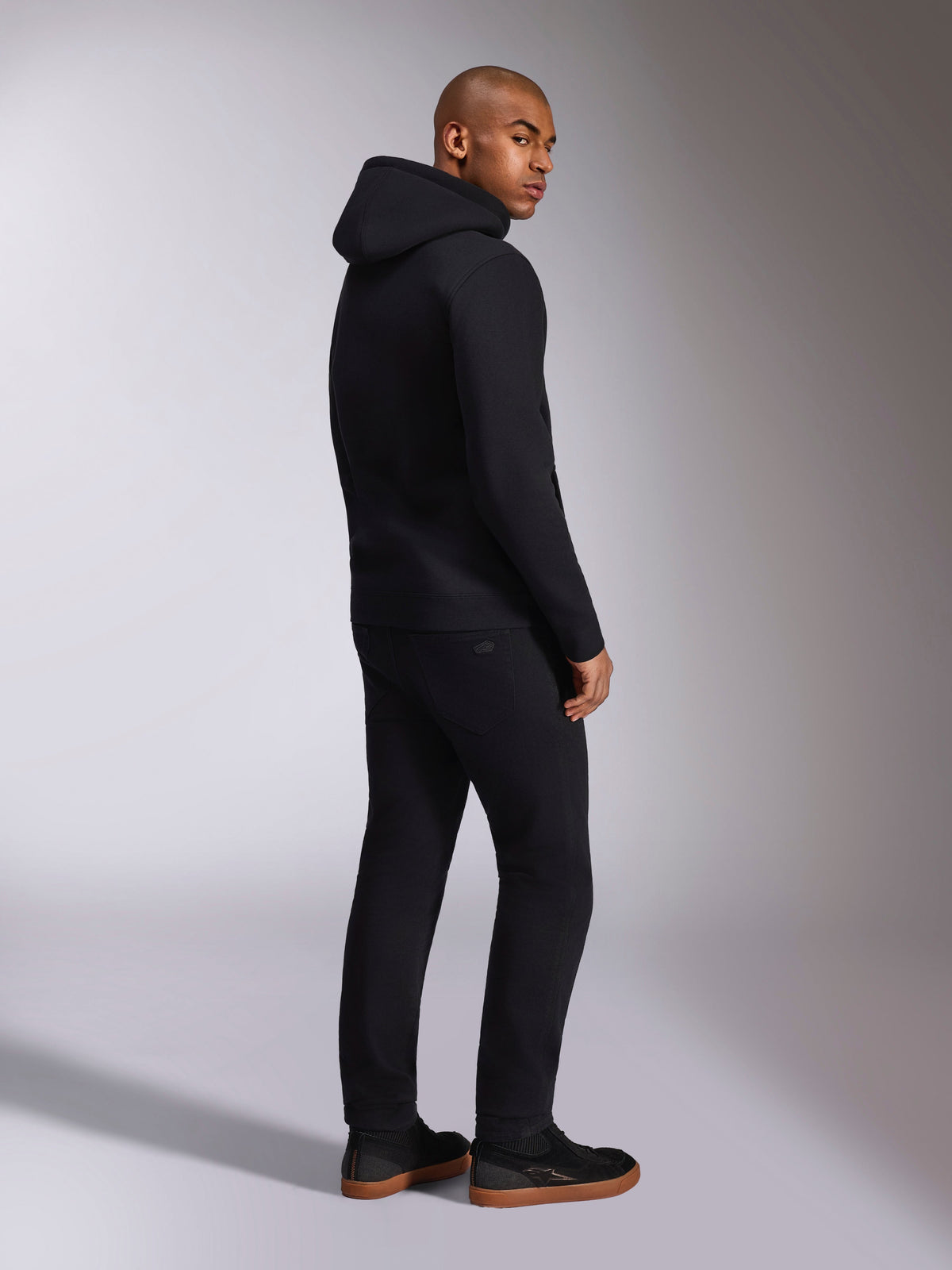 Alpinestars Ellipsoid Hoodie, ropa urbana casual negra, vista trasera en modelo masculino, sudadera de forro polar con capucha, puños y dobladillo de canalé, combinada con pantalones negros y zapatillas, diseño moderno inspirado en el automovilismo