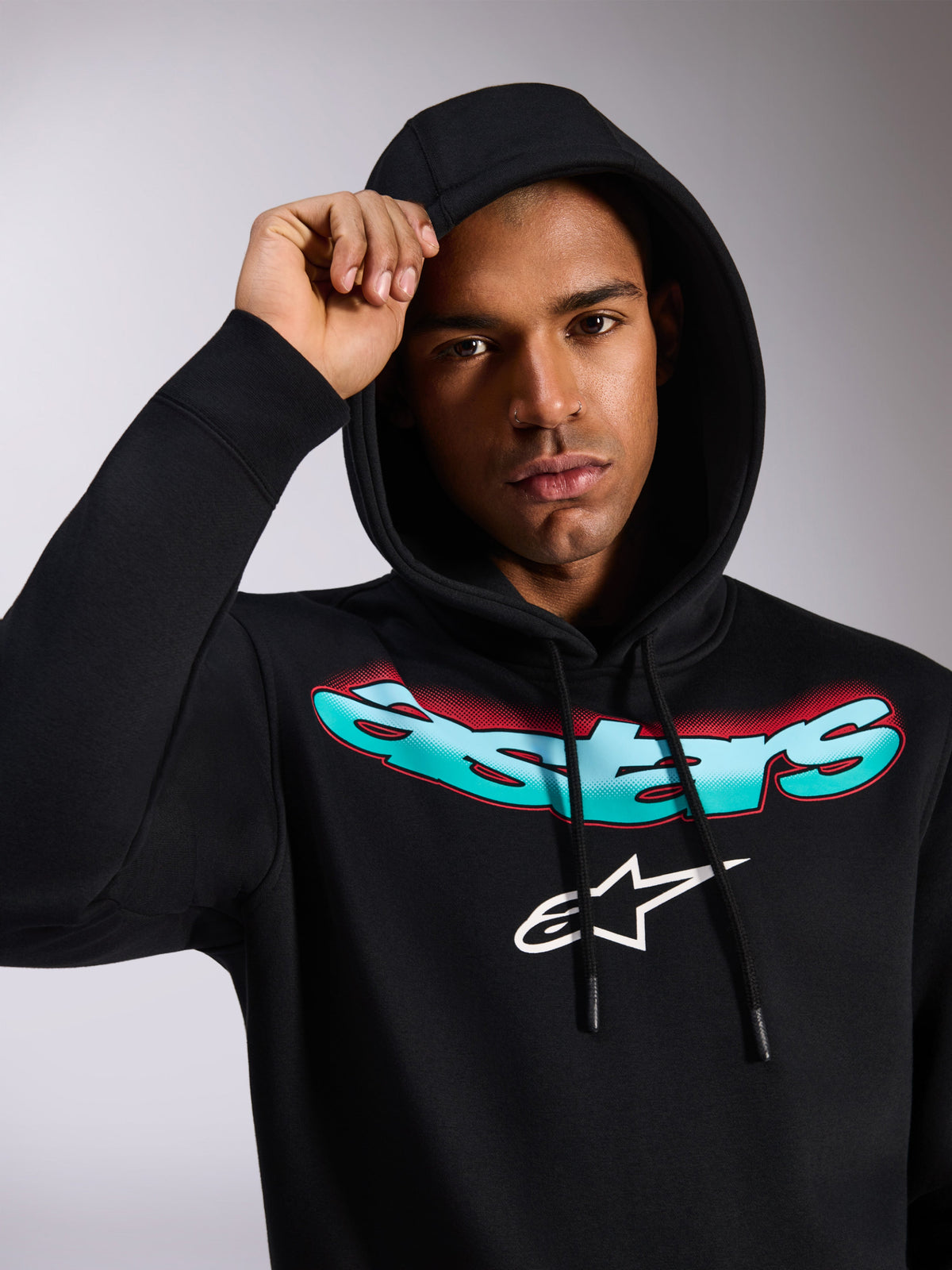 Alpinestars Ellipsoid Hoodie, sudadera negra con capucha, con la palabra Astars estilizada en degradado de color verde azulado y rojo y logotipo blanco en el pecho, lucida por un modelo con la capucha puesta, ropa de calle informal inspirada en el automovilismo