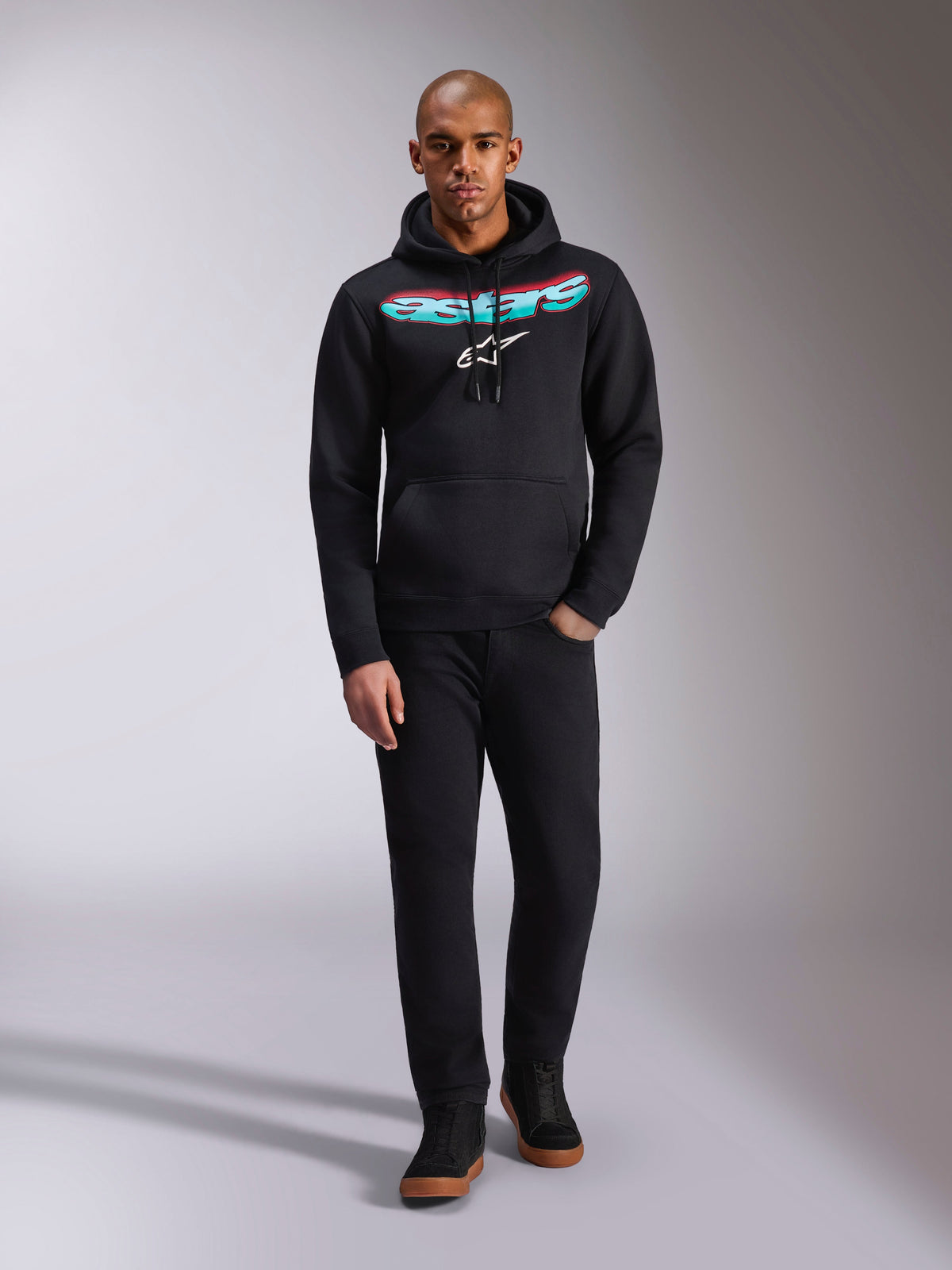 Sudadera con capucha Alpinestars Ellipsoid, negra, sudadera de felpa con la palabra Astars curva en degradado rojo y verde azulado y logotipo de estrella blanca, usada por un modelo con pantalones negros y zapatillas, estilo urbano casual de motorsport