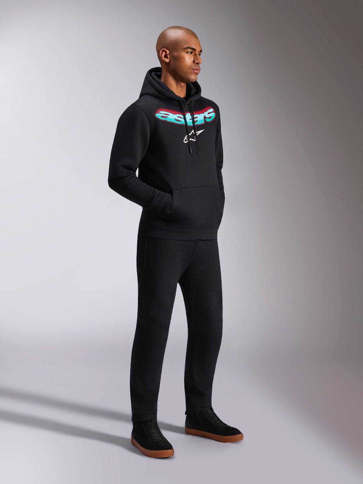 Sudadera Alpinestars Ellipsoid Hoodie, sudadera negra de forro polar con capucha, con el nombre Astars curvado en degradado verde azulado y rojo y logotipo de estrella blanca, lucida por un modelo con pantalones negros, ropa urbana de estilo casual inspirado en el motor