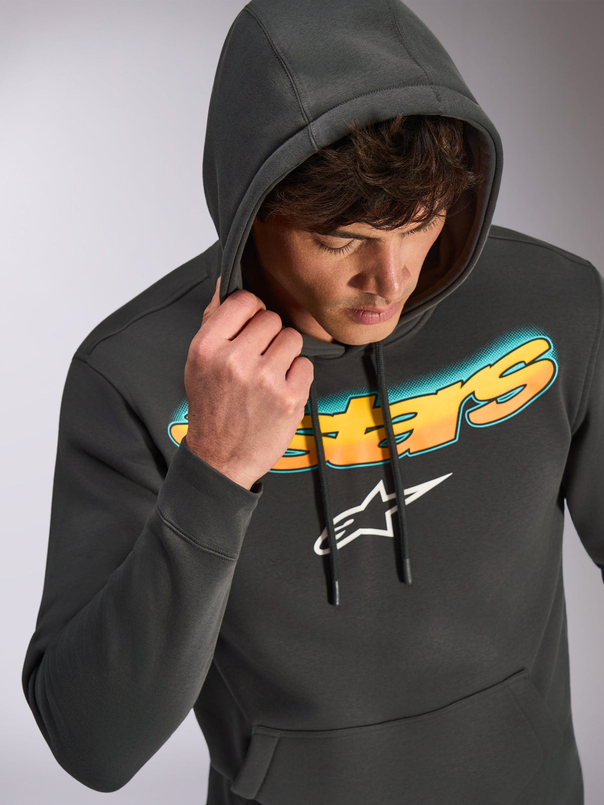 Sudadera con capucha Alpinestars Ellipsoid, gris Gunmetal, sudadera informal tipo pullover con la palabra Astars en degradado de cian a naranja y logotipo blanco, lucida por un modelo con la capucha puesta, ropa urbana inspirada en el automovilismo