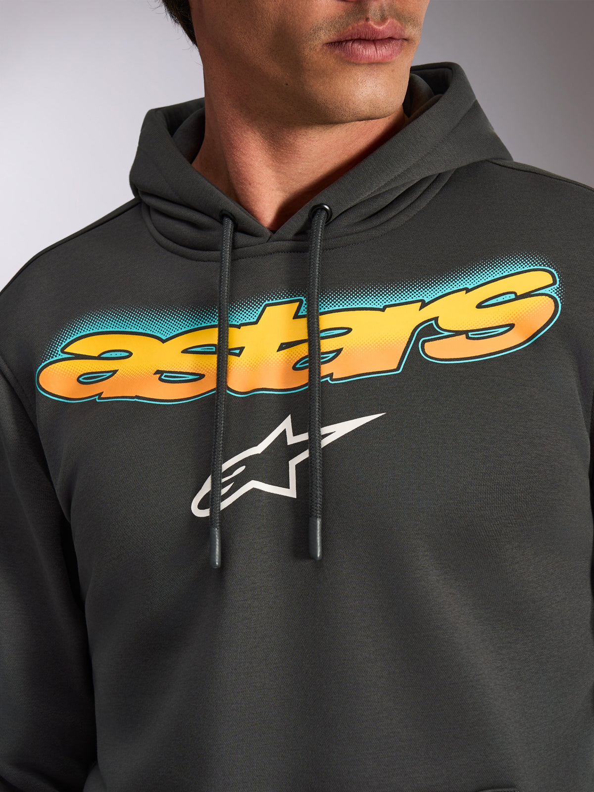 Sudadera Alpinestars Ellipsoid, gris Gunmetal, primer plano del pecho con la palabra 'astars' curvada en degradado de amarillo a naranja y detalles en verde azulado, logotipo de la estrella Alpinestars en blanco, forro polar casual inspirado en el motor