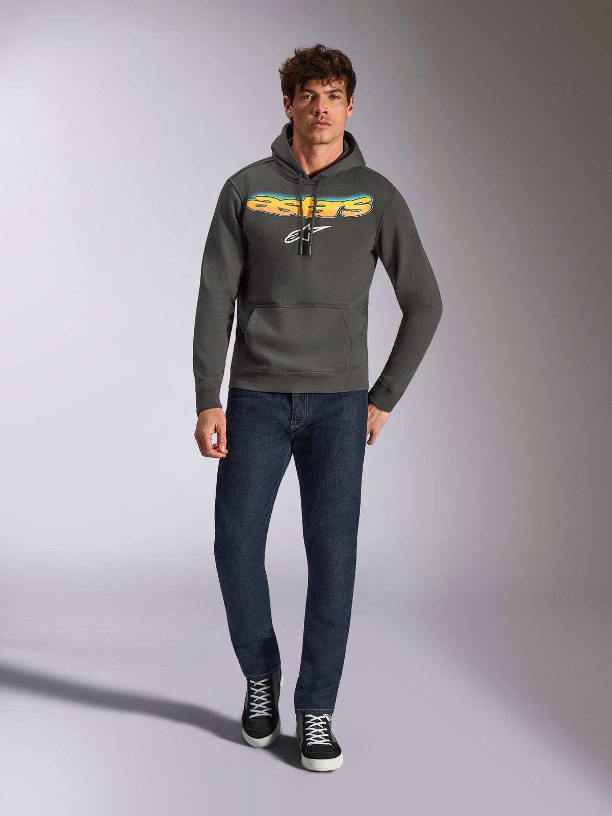 Sudadera con capucha Alpinestars Ellipsoid, ropa deportiva casual, gris gunmetal con logotipo Astars en degradado naranja y amarillo y símbolo blanco, capucha con cordón, usada por un modelo masculino con vaqueros azul oscuro y zapatillas negras, estilo streetwear moderno inspirado en el motor