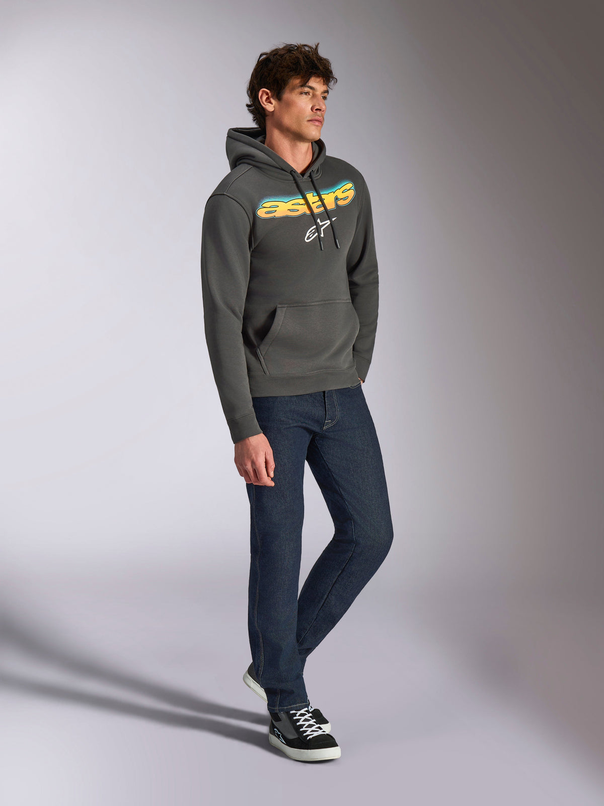 Sudadera con capucha Alpinestars Ellipsoid, forro polar gris gunmetal, palabra Astars curvada en el frente con degradado de azul y amarillo y logotipo blanco, combinada con vaqueros oscuros y zapatillas negras, ropa urbana casual