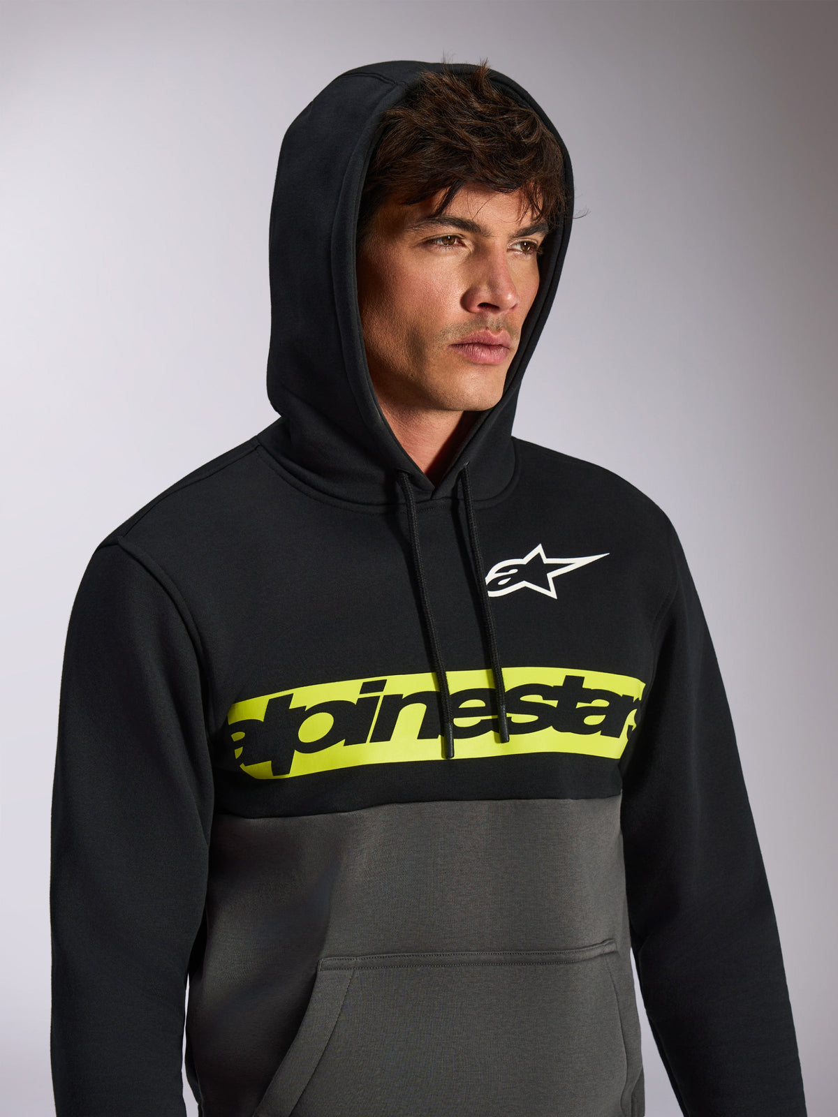 Sudadera con capucha Alpinestars Parallel, estilo streetwear casual con bloques de color, parte superior negra y parte inferior gris plomo, banda amarilla neón en el pecho con el logo de Alpinestars en negro, logo Astars blanco, jersey con capucha y cordón
