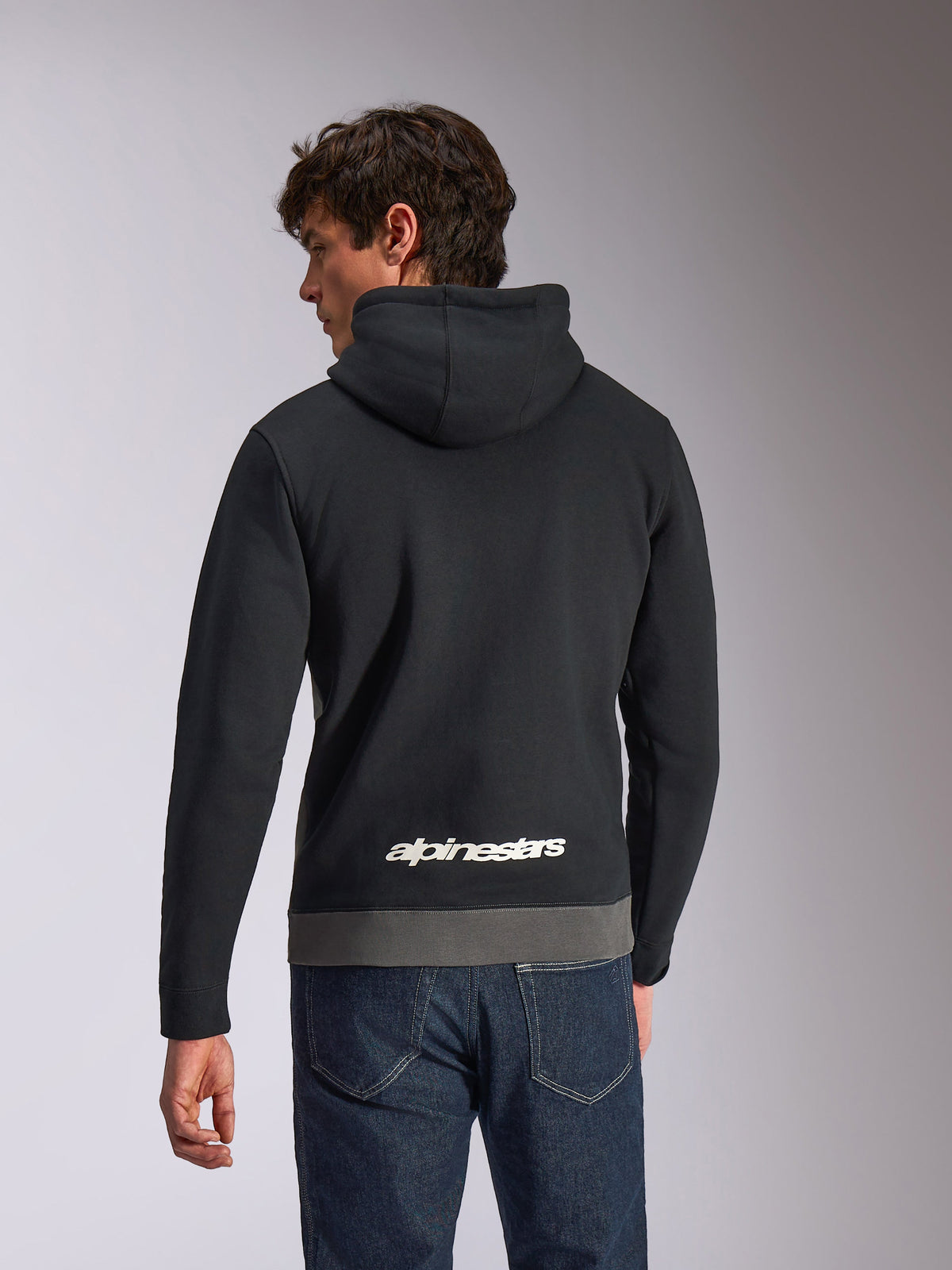 Sudadera Alpinestars Parallel, sudadera deportiva casual, negro y gunmetal, vista trasera con un logotipo blanco de Alpinestars en la parte inferior de la espalda, diseño de bloques de color con dobladillo gris, combinada con vaqueros de denim oscuro