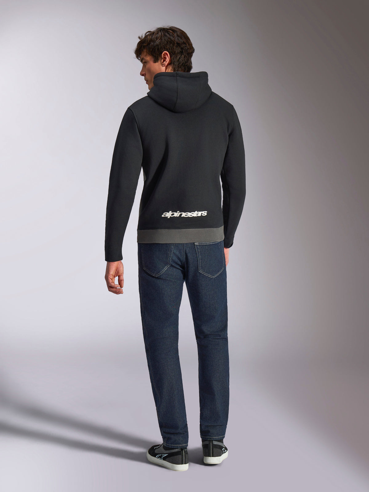 Sudadera con capucha Alpinestars Parallel, vista trasera de un hombre con un forro polar de tipo pullover en bloques de color negro y gunmetal, logotipo de Alpinestars en blanco en la parte inferior de la espalda, combinado con vaqueros azul oscuro, ropa urbana casual inspirada en el motociclismo
