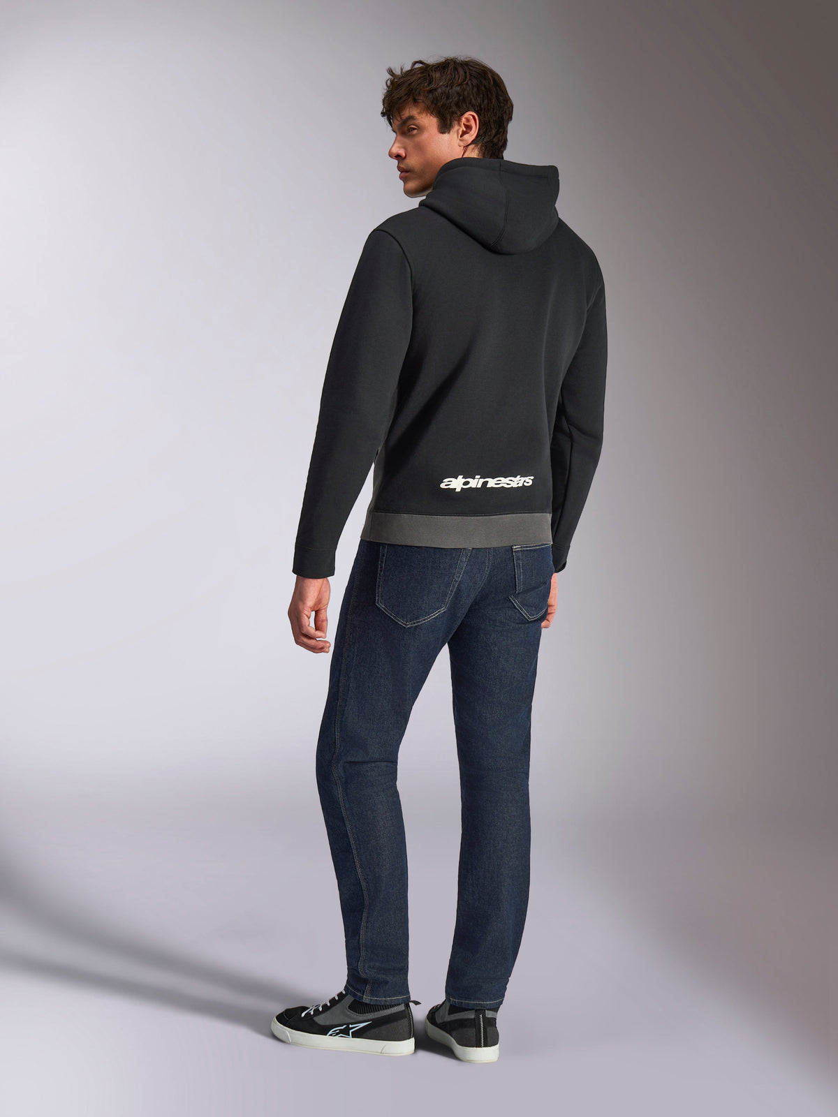 Sudadera Parallel de Alpinestars, vista posterior del modelo con el jersey en negro y gris plomo, con el logotipo blanco de Alpinestars en la parte inferior de la espalda, combinado con vaqueros azul oscuro, estilo urbano casual inspirado en el motor