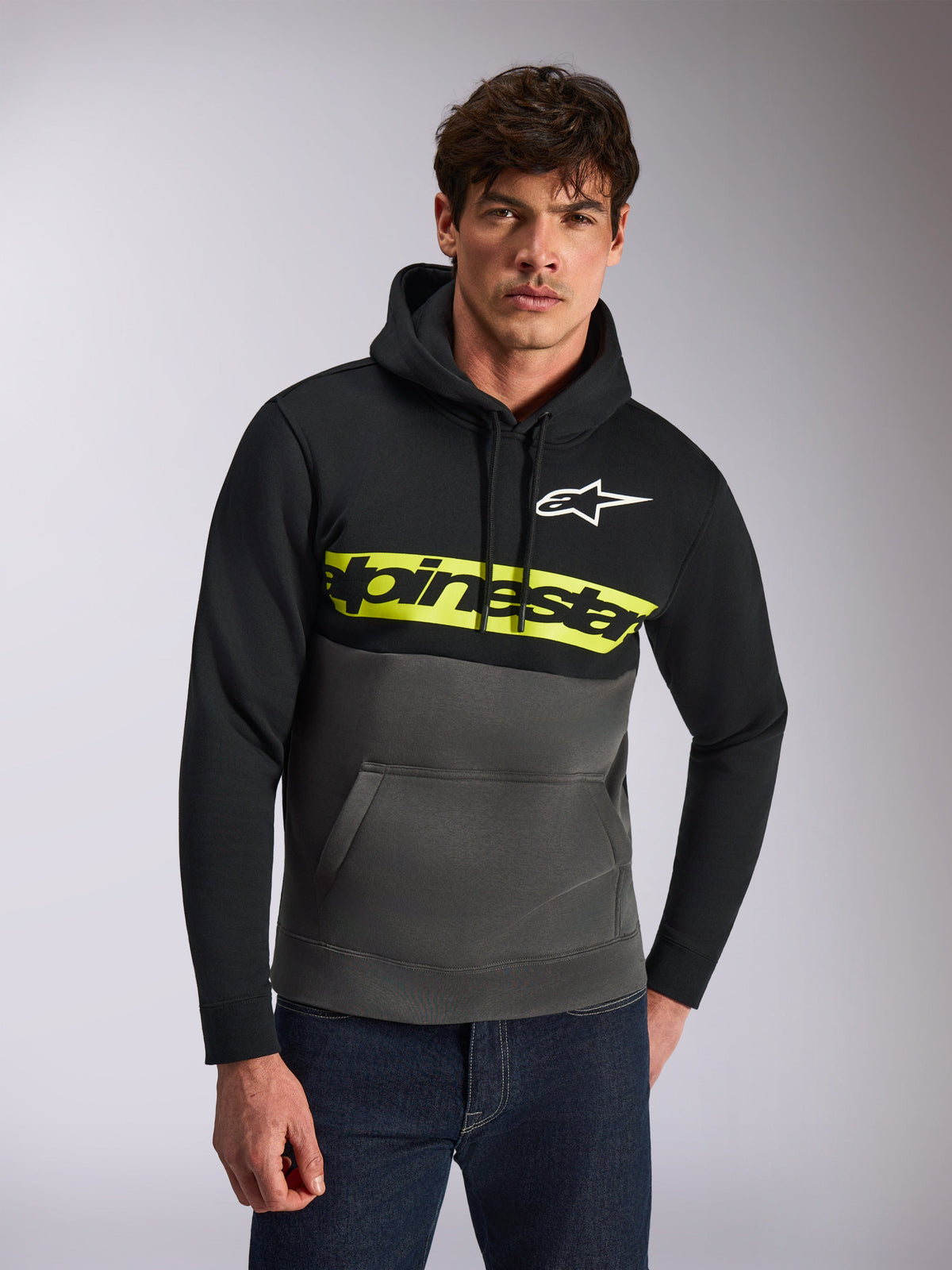 Sudadera con capucha Alpinestars Parallel, estilo streetwear casual, bloques de color negro y gris gunmetal, franja amarilla brillante en el pecho con el logotipo de Alpinestars, logotipo Astars blanco, usada por un modelo con vaqueros azules, diseño moderno inspirado en el motorsport