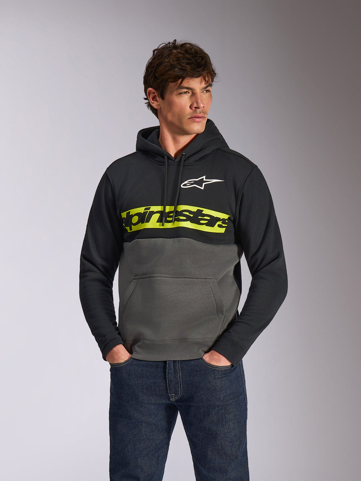 Sudadera con capucha Alpinestars Parallel, sudadera casual con bloques de color, negro y gris plomo con una franja amarilla vibrante en el pecho, logotipo de Alpinestars y logo de estrella blanca, usada por un modelo masculino con vaqueros azul oscuro
