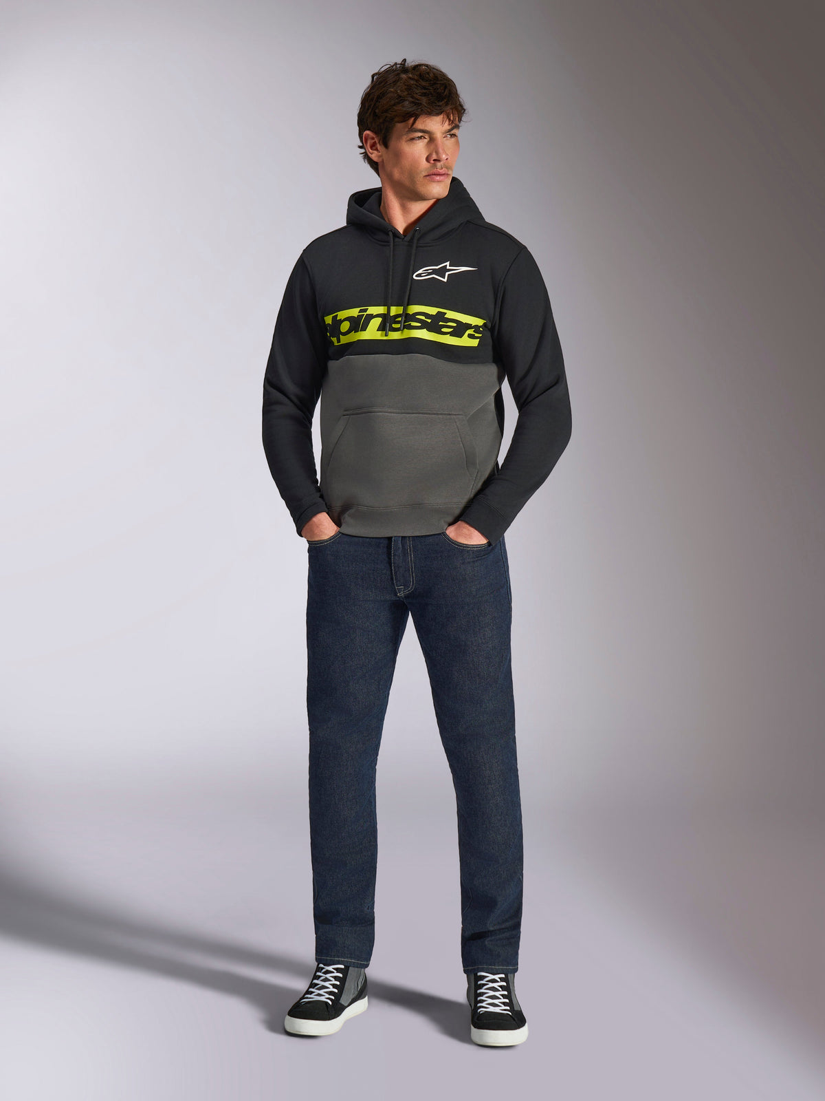 Sudadera con capucha Alpinestars Parallel, diseño de bloques de color en negro y gris plomo, logotipo de Alpinestars en amarillo brillante y logotipo de estrella blanca en el pecho, usada por un modelo masculino con vaqueros azul oscuro y zapatillas negras, estilo streetwear casual de motor