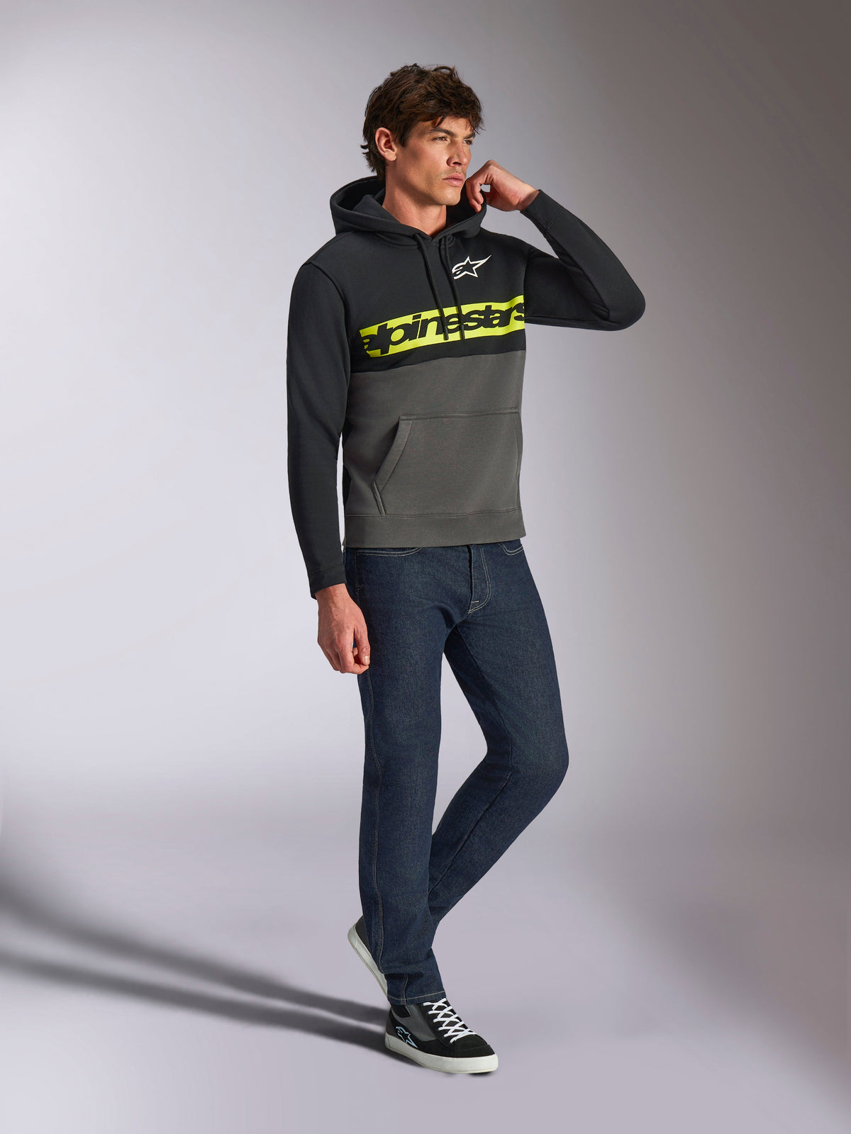 Sudadera con capucha Alpinestars Parallel, estilo streetwear casual, bloques de color en negro y gris plomo, logotipo Alpinestars en amarillo neón en el pecho, logotipo Astars en blanco, capucha con cordón, combinada con vaqueros oscuros y zapatillas