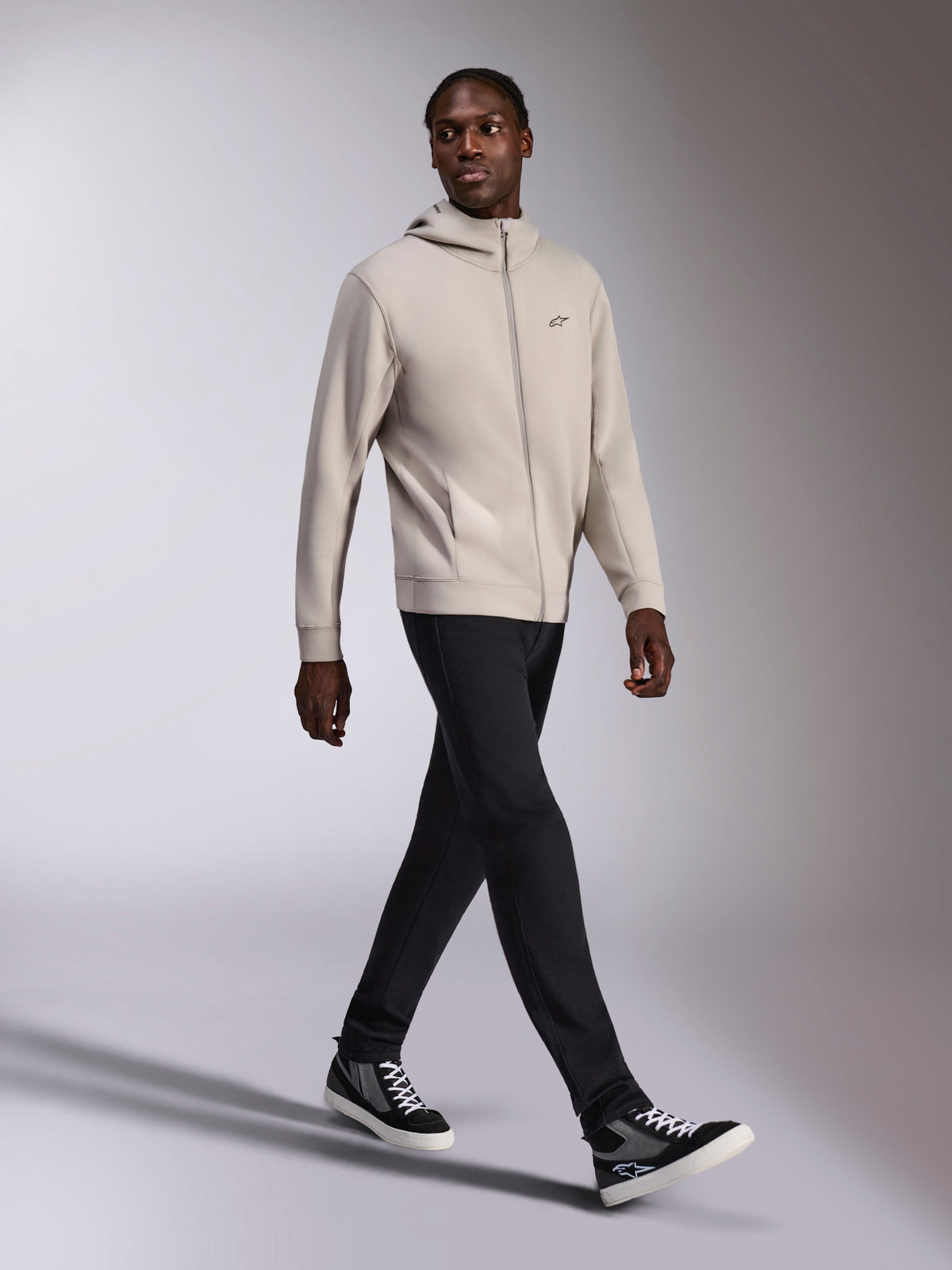 Alpinestars Cypher Hoodie, Cement gray full-zip fleece, high neck hood, small Astars chest logo, worn by a model walking, paired with black pants and black and gray sneakers, casual motorsport-inspired sportswear.