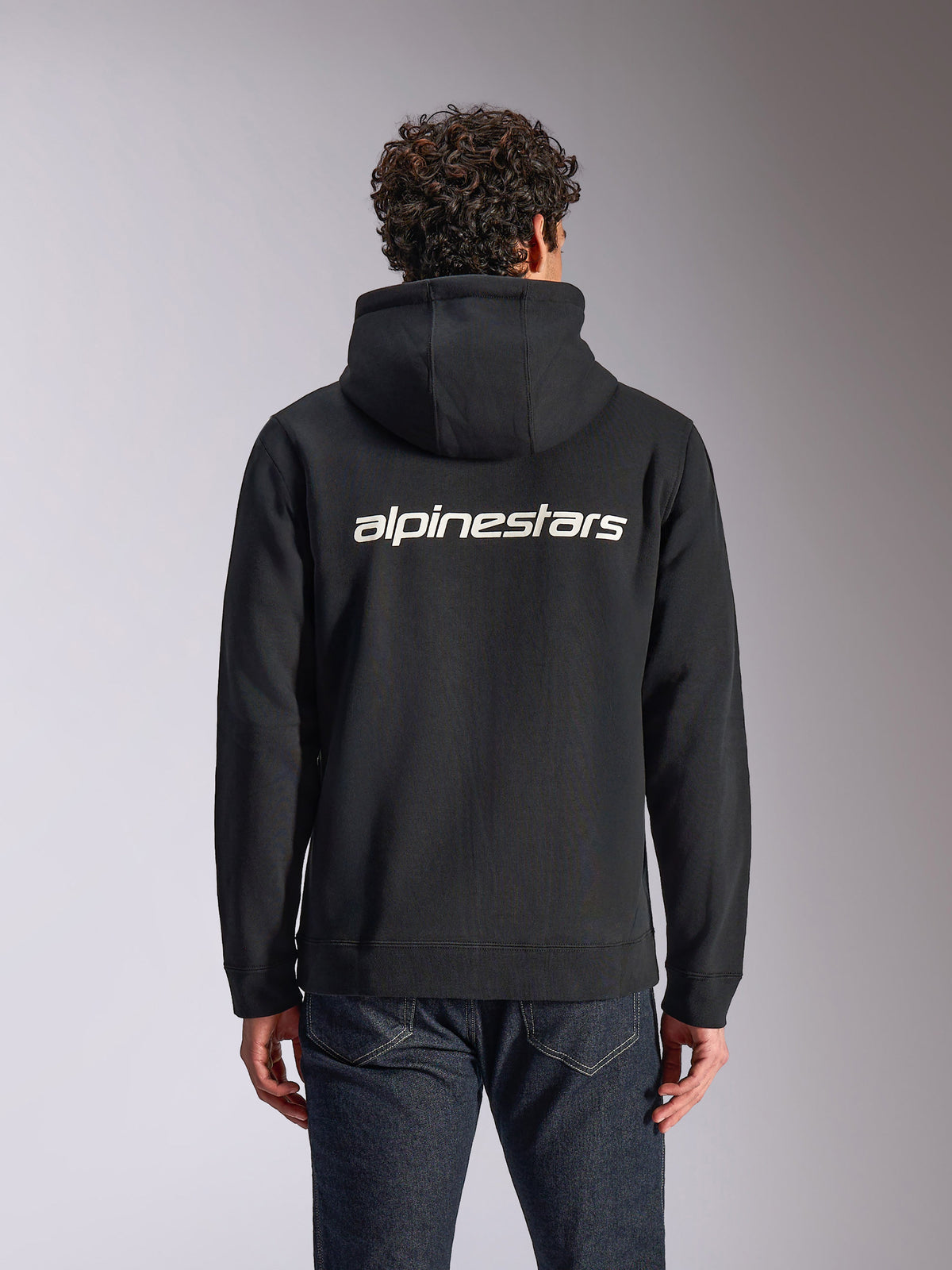 Alpinestars Transverse Hoodie, casual streetwear, black with white Alpinestars word logo on the back, zip-front hooded fleece, long sleeves, worn by a model with dark denim jeans, modern motorsport-inspired design