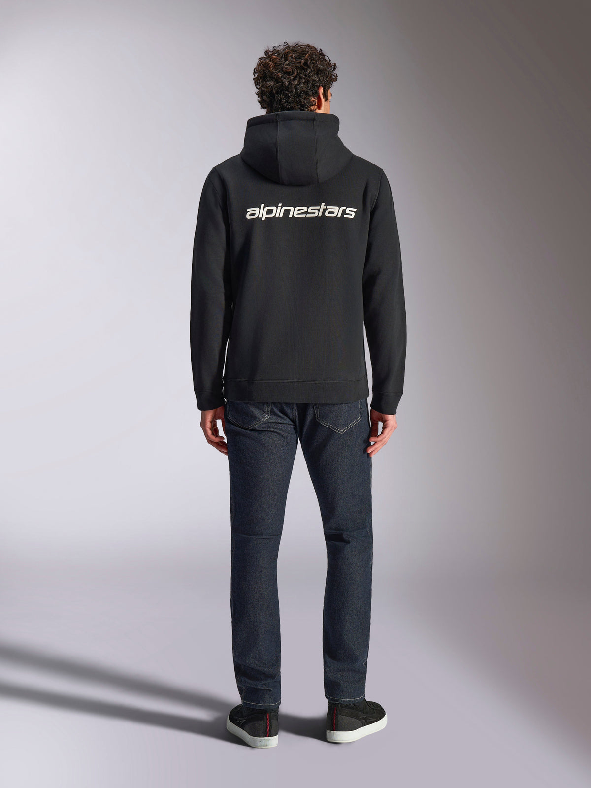 Alpinestars Transverse Hoodie, casual motorsport streetwear, Black with white Alpinestars word logo on the back, long sleeves, ribbed hem and cuffs, worn from behind with dark denim jeans