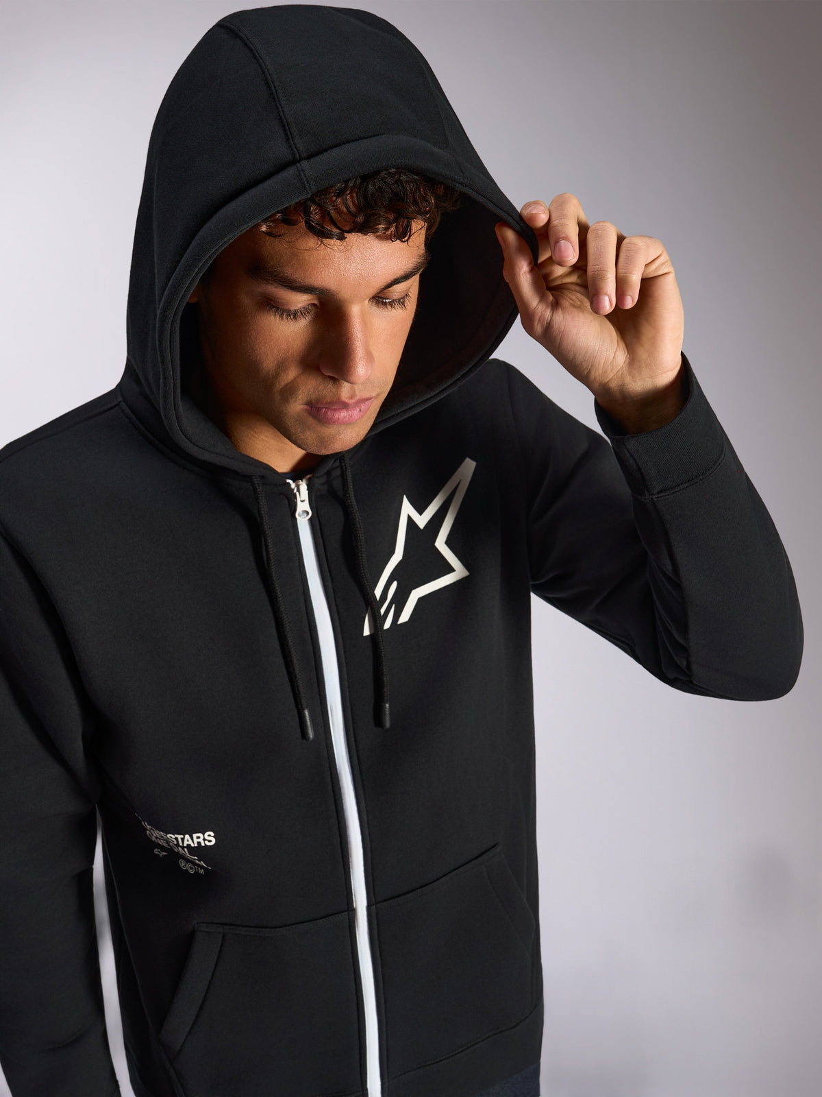 Alpinestars Transverse Hoodie, black zip-front hoodie, white Astars logo on chest, white zipper, drawstring hood worn up, casual motorsport-inspired streetwear