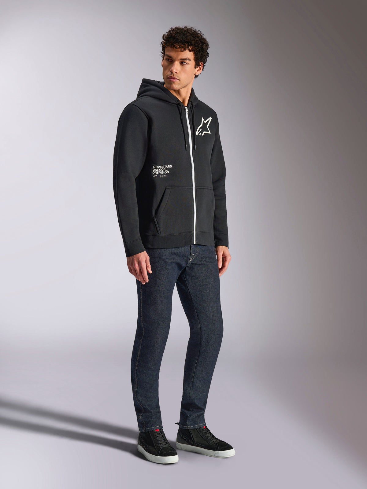 Alpinestars Transverse Hoodie, black zip-up hoodie with a contrast white zipper, featuring a white star logo on the chest and 'One Goal' branding on the pocket, worn by a model with dark jeans and black sneakers, casual motorsport-inspired streetwear.