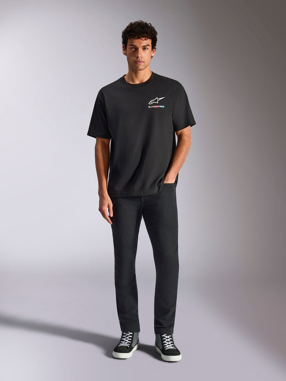 Alpinestars Wild Oversized CSF Tee, camiseta de manga corta, negra, lucida por un modelo con pantalones negros y zapatillas, con el logo Astars blanco y un diseño de marca multicolor vibrante, estilo casual streetwear de automovilismo