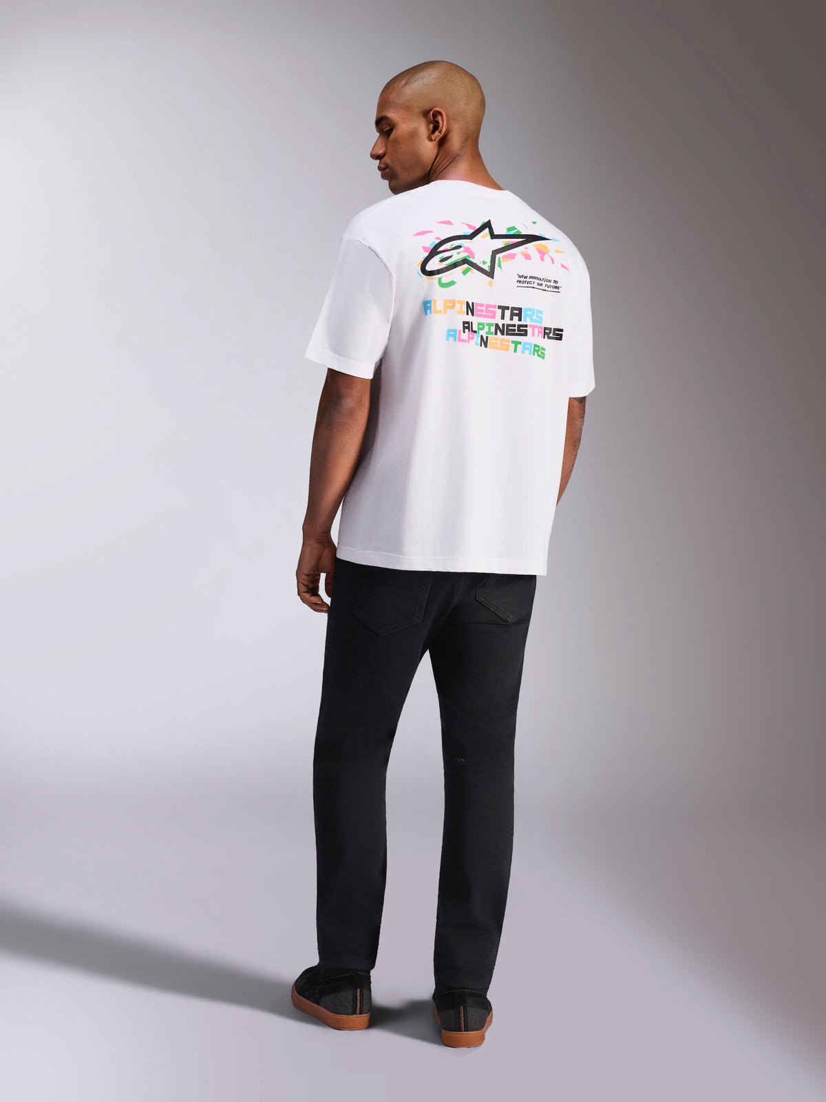 Alpinestars Wild Oversized CSF Tee, camiseta de manga corta, blanca, vista trasera del modelo usando la camiseta de corte grande con el vibrante logotipo Astars multicolor y texto apilado en la espalda, ropa urbana casual inspirada en el motorsport