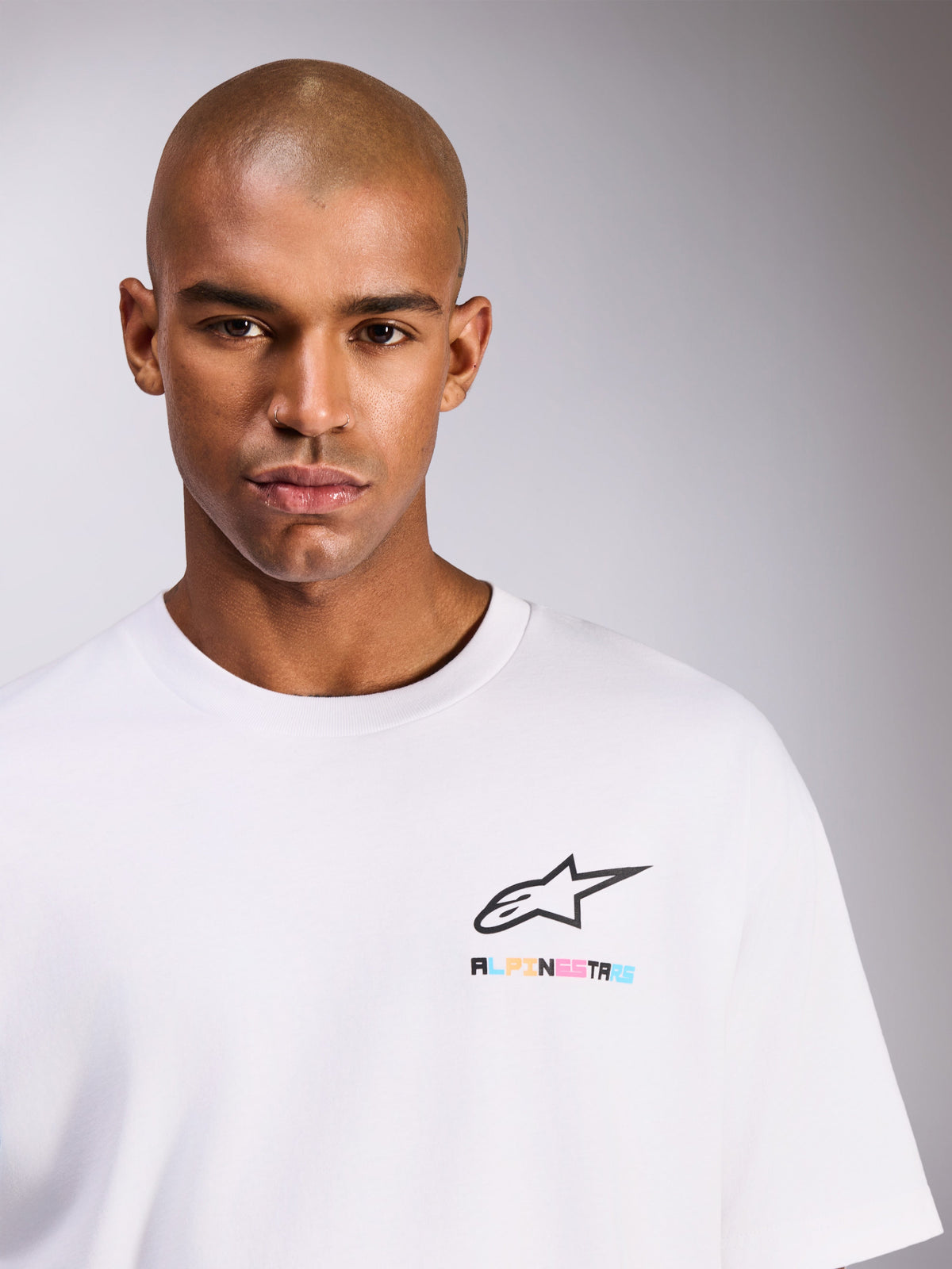 Modelo masculino con la camiseta Alpinestars Wild Oversized CSF, camiseta de manga corta en color blanco, con el logo Astars negro y letras multicolor vibrantes en el pecho, estilo casual streetwear