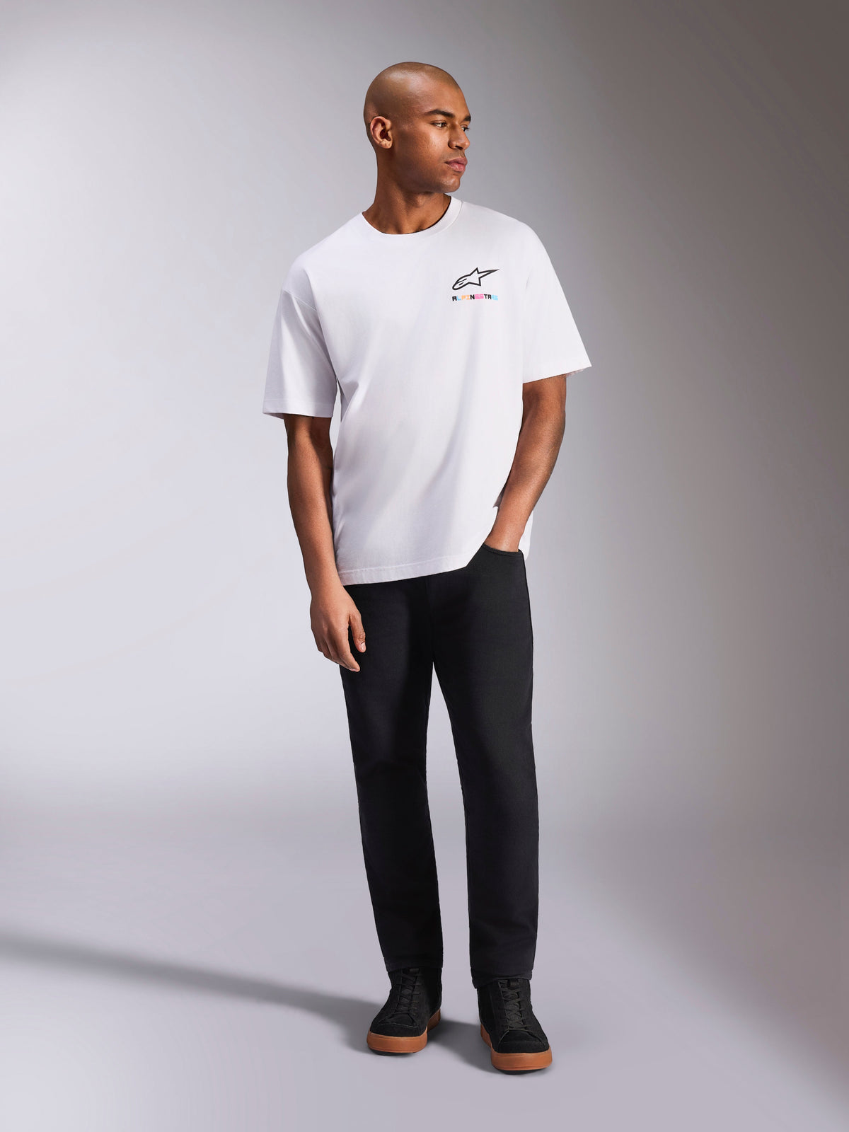 Camiseta Alpinestars Wild Oversized CSF, camiseta de manga corta blanca, logotipo de Astars multicolor vibrante en el pecho, combinada con pantalones negros y zapatillas, ropa casual de estilo motorsport