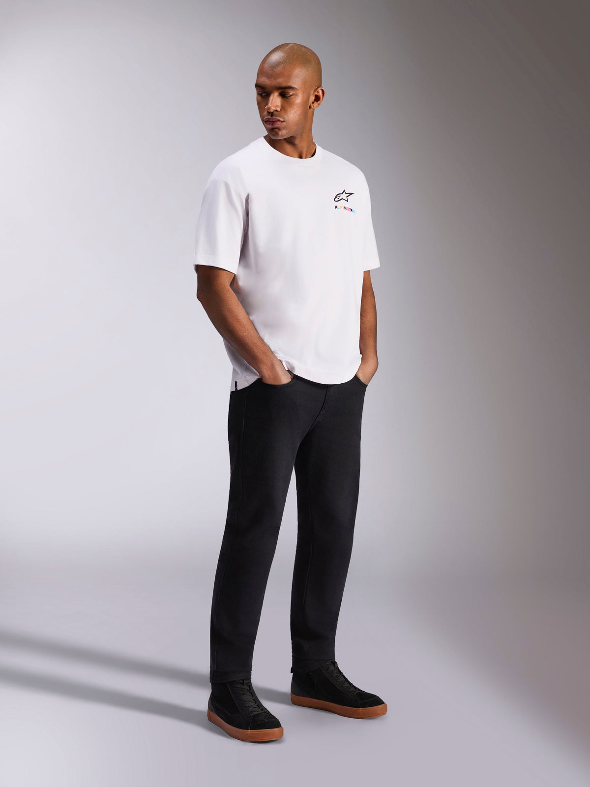 Camiseta Alpinestars Wild Oversized CSF, camiseta de manga corta, blanca, con un pequeño logo Astars multicolor en el pecho, llevada por un modelo con pantalones negros y zapatillas negras, ropa urbana casual inspirada en el automovilismo