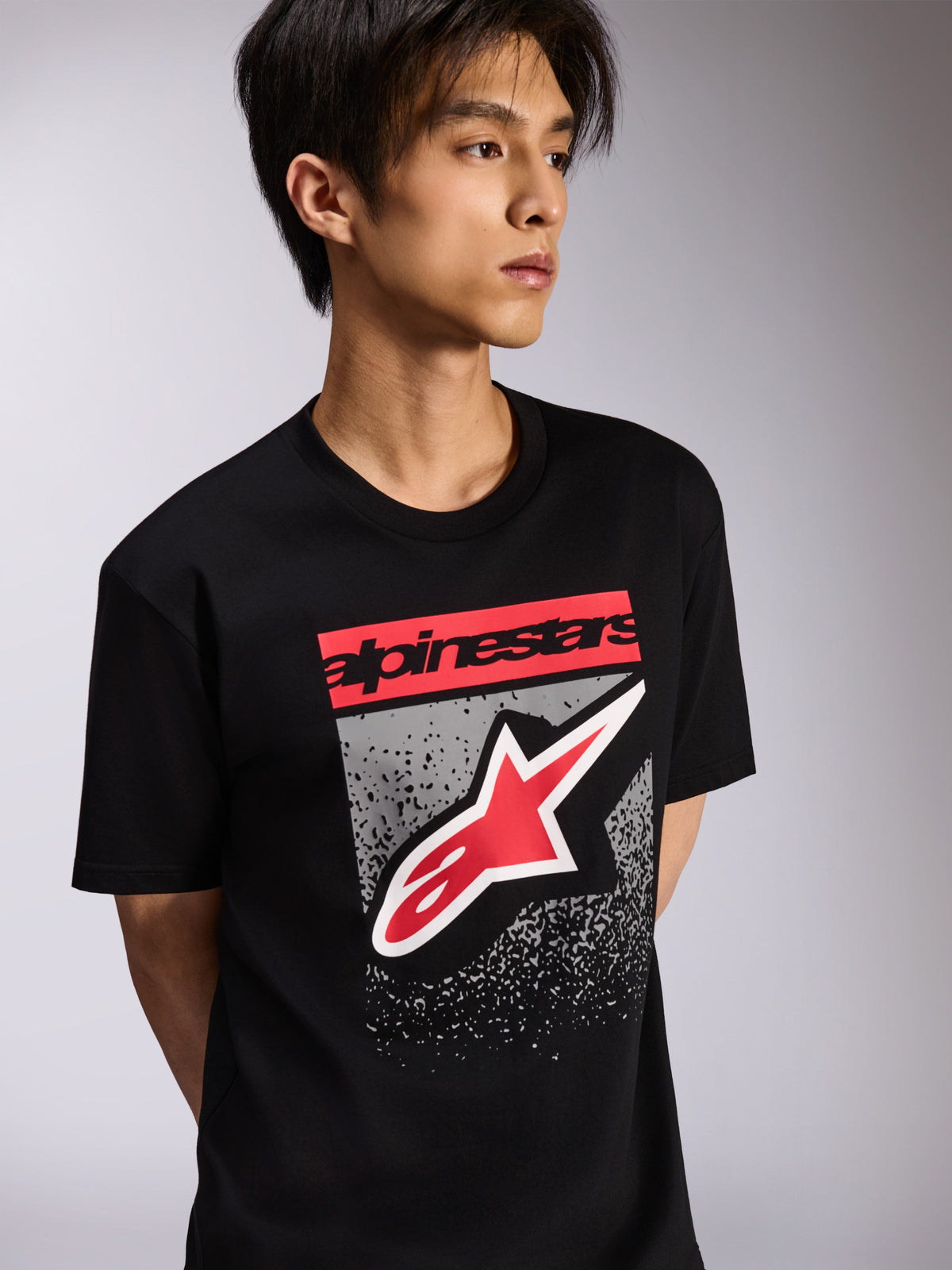 Alpinestars Noka CSF Tee, camiseta de manga corta, negra, con un bloque de marca rojo y un gran logotipo Astars en rojo y blanco sobre un fondo gris texturizado, estilo casual de automovilismo
