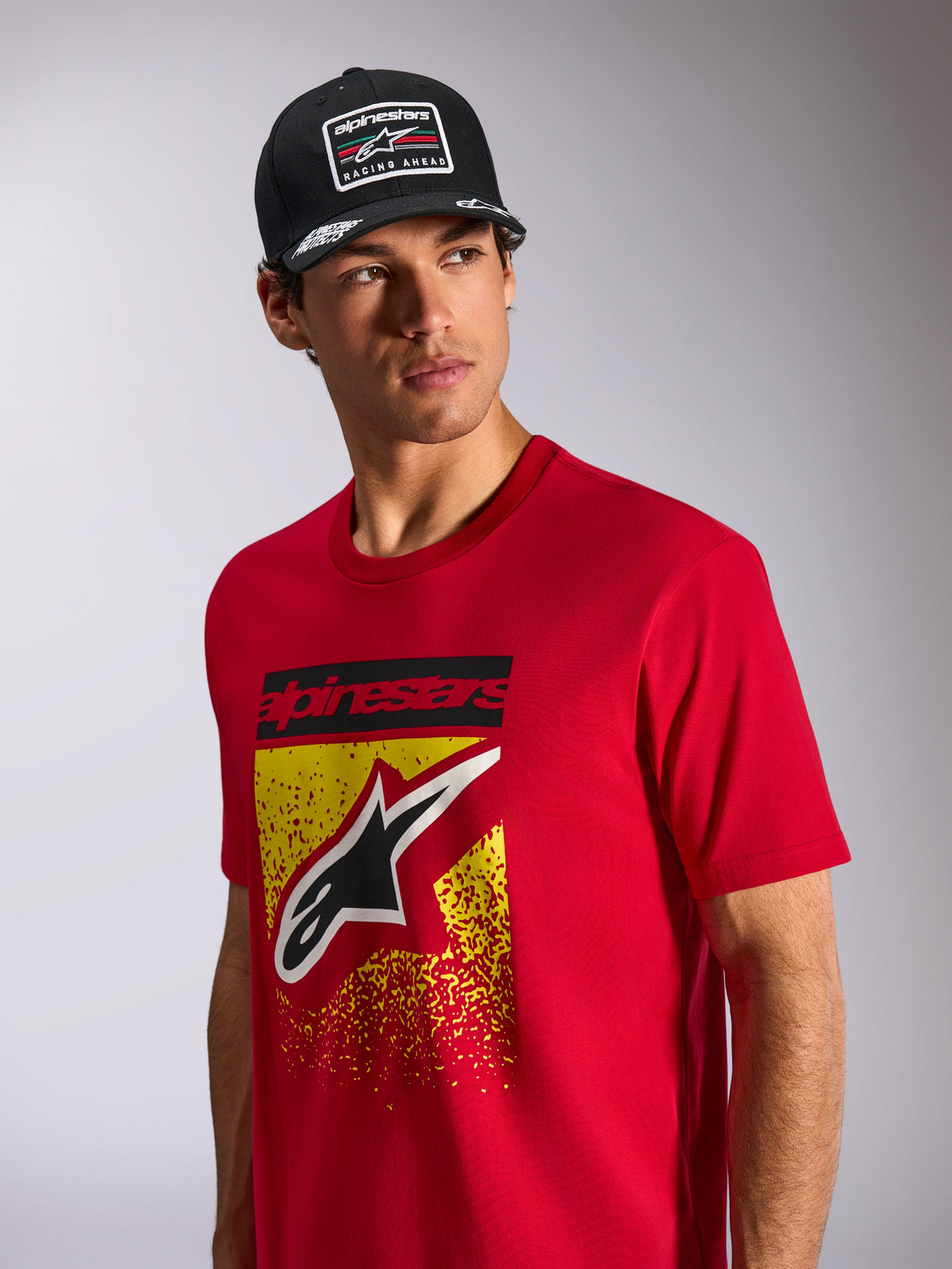 Alpinestars Noka CSF Tee, Short Sleeve T-Shirt, Red, featuring a front Astars logo with yellow and red MX gradation, worn with a black Alpinestars 'Racing Ahead' cap, casual motorsport streetwear
