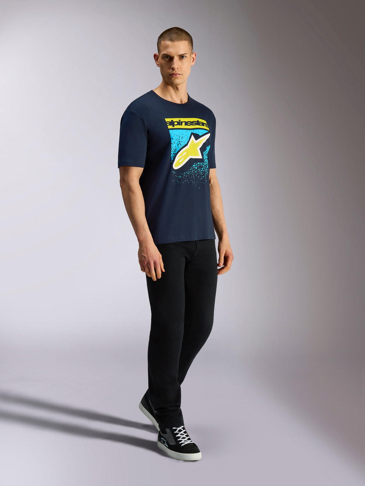 Alpinestars Noka CSF Tee, camiseta de manga corta, azul marino, logotipo Astars amarillo con gráfico de degradado MX azul, modelo masculino con camiseta azul marino, pantalones negros y zapatillas, ropa urbana casual de estilo motorsport