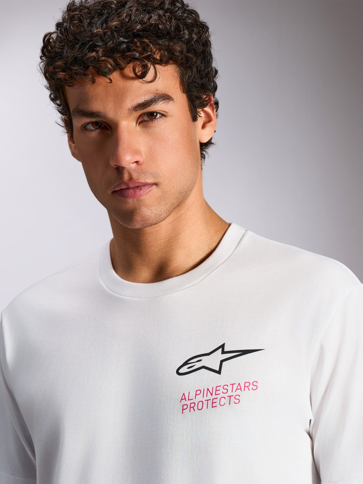 Camiseta Alpinestars Pop Helmet CSF, camiseta de manga corta blanca, logotipo de Astars en negro y texto 'ALPINESTARS PROTECTS' en rosa en el pecho, ropa urbana casual de estilo motorsport