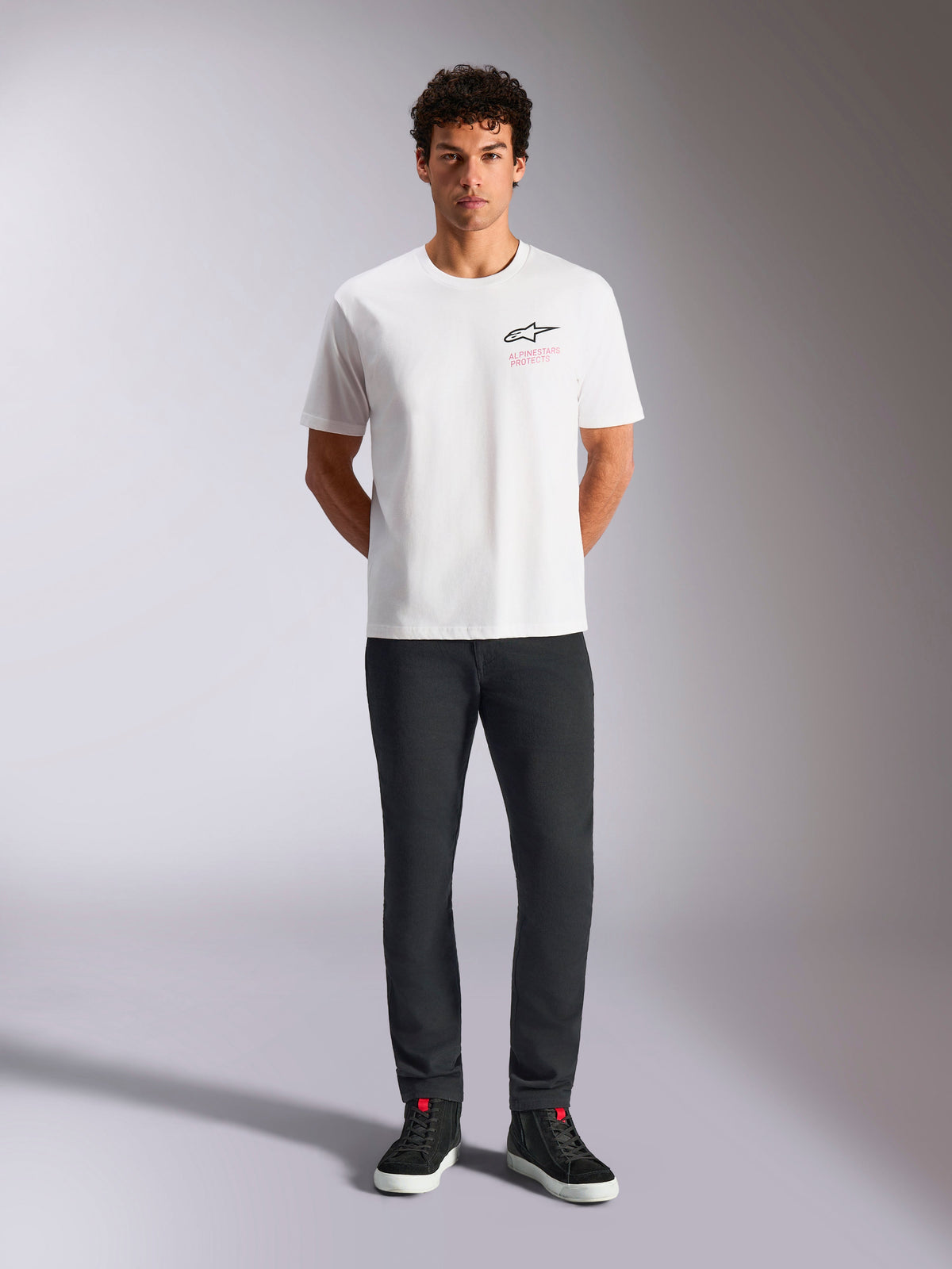Alpinestars Pop Helmet CSF Tee, camiseta de manga corta, blanca, lucida por un modelo con pantalones negros y zapatillas de caña alta negras, logotipo frontal de Astars con texto 'Alpinestars Protects' en rosa, estilo streetwear casual de automovilismo